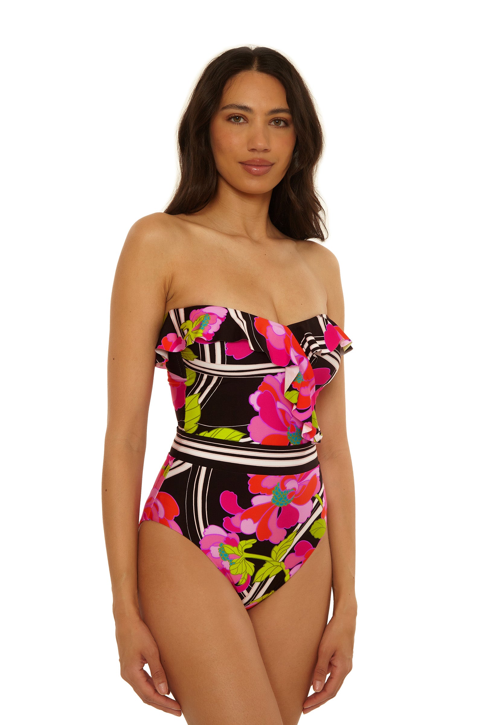 A woman models the Bandeau One-Piece Swimsuit - Bougainvillea, featuring a strapless, ruffled design with a vibrant pink, green, and white floral pattern on black, set against a plain white background.