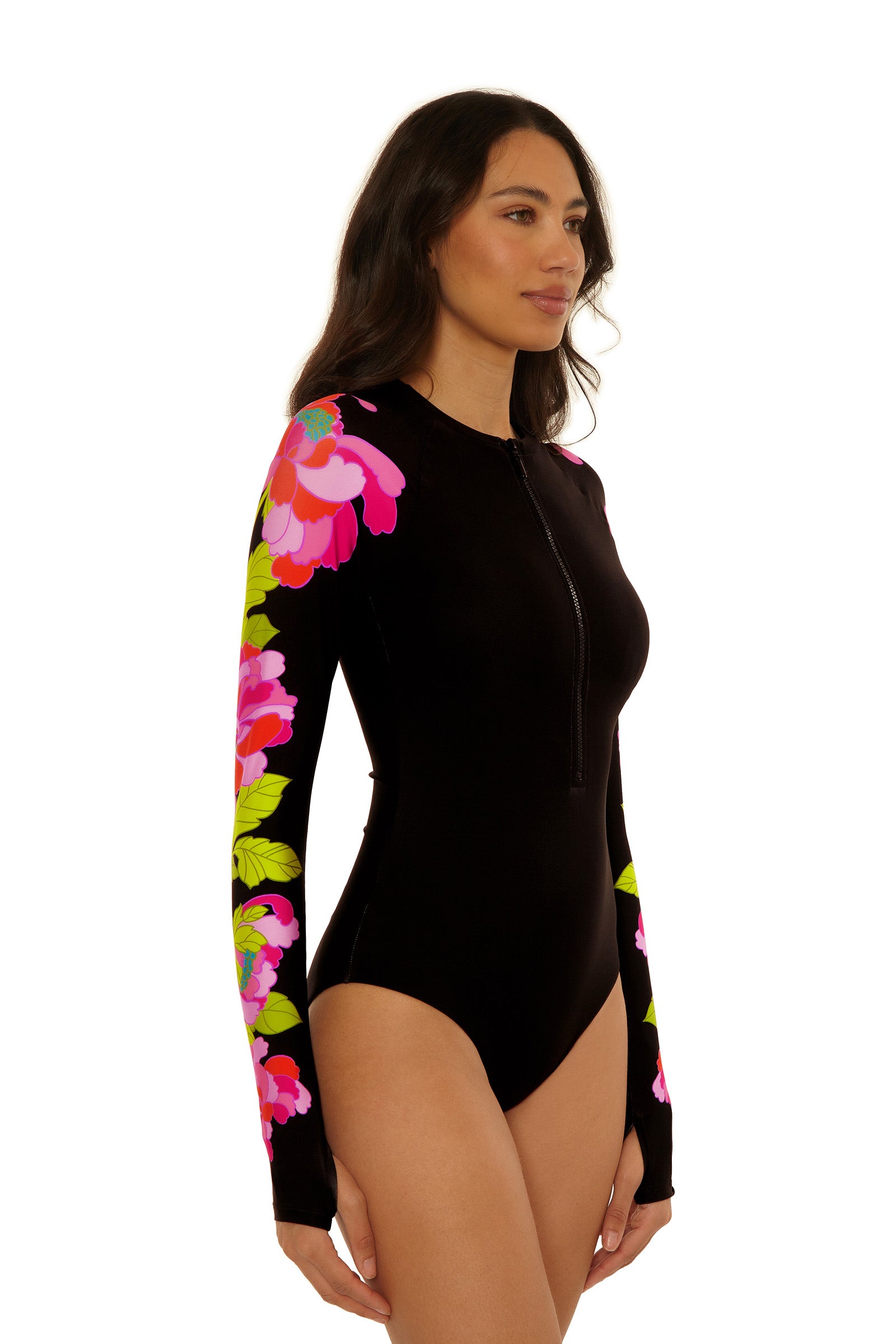 A woman models the Long-Sleeve Zip-Up Paddlesuit – Bougainvillea, featuring a black body, front zipper, and vibrant pink, red, and green bougainvillea floral sleeves as she stands slightly sideways.