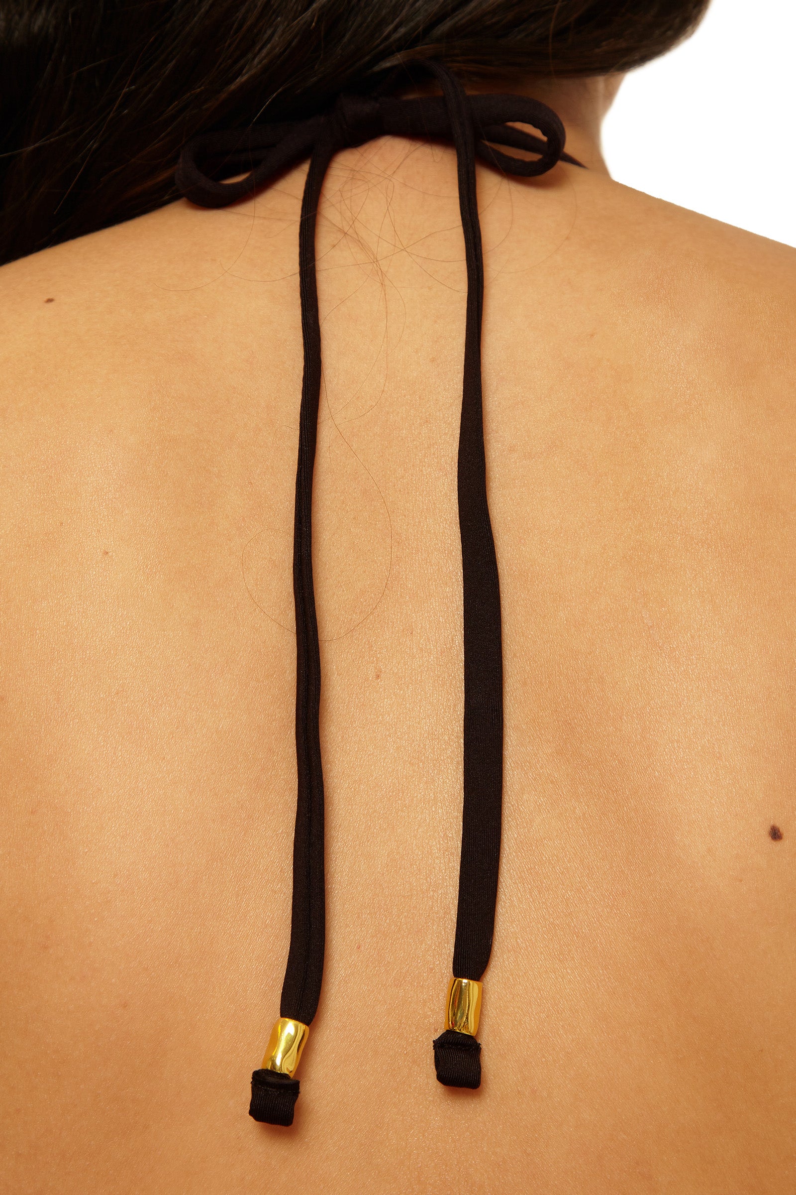 Close-up of a persons upper back wearing the Reversible Halter Swim Top - Bougainvillea, featuring black halter straps tied in a bow at the neck with gold-colored metal tips. The background is plain white.