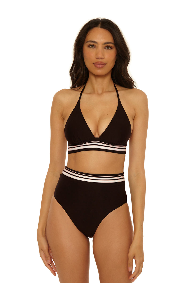 A woman stands facing forward in the High-Waist Swim Bottom - Bougainvillea, a black bikini with a halter top and high-waisted bottoms. Both pieces have white and pink horizontal stripes on the bands. The background is plain white.