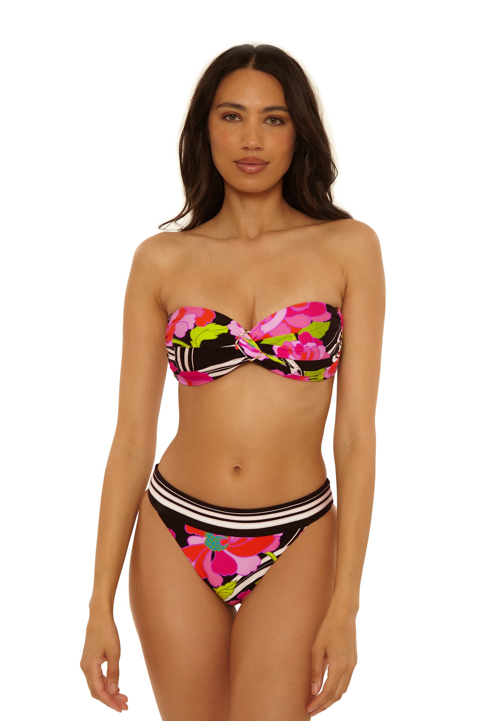 A woman wears the Reversible Hipster Swim Bottom in Bougainvillea Swim Knit with a strapless, twisted bandeau bikini top, featuring a bold pink, red, green, and black floral pattern, standing against a plain white background.