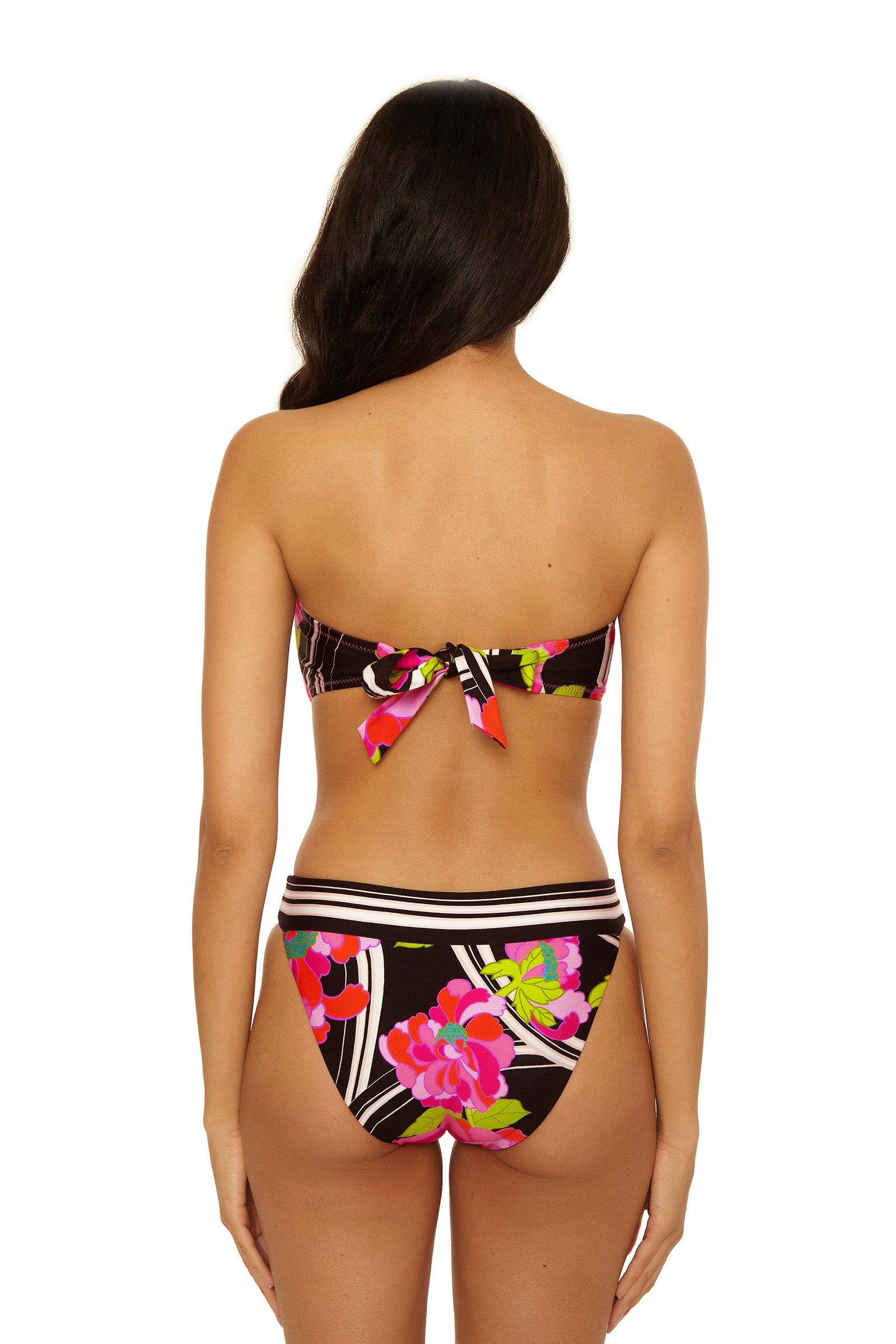 A woman with long dark hair is seen from the back wearing the Reversible Hipster Swim Bottom - Bougainvillea, featuring a bright pink, green, and white floral pattern on black. The bikini top is tied in a bow at the back.