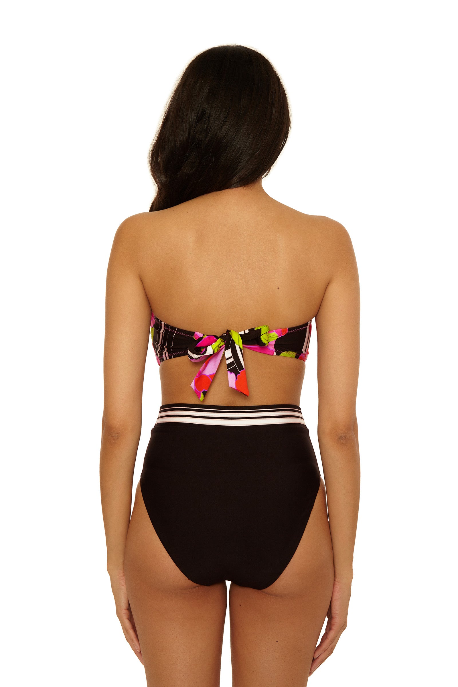 A woman with long dark hair stands facing away, wearing the Twist Bandeau Swim Top - Bougainvillea featuring a colorful back bow and high-waisted black bikini bottoms with a white-striped waistband.