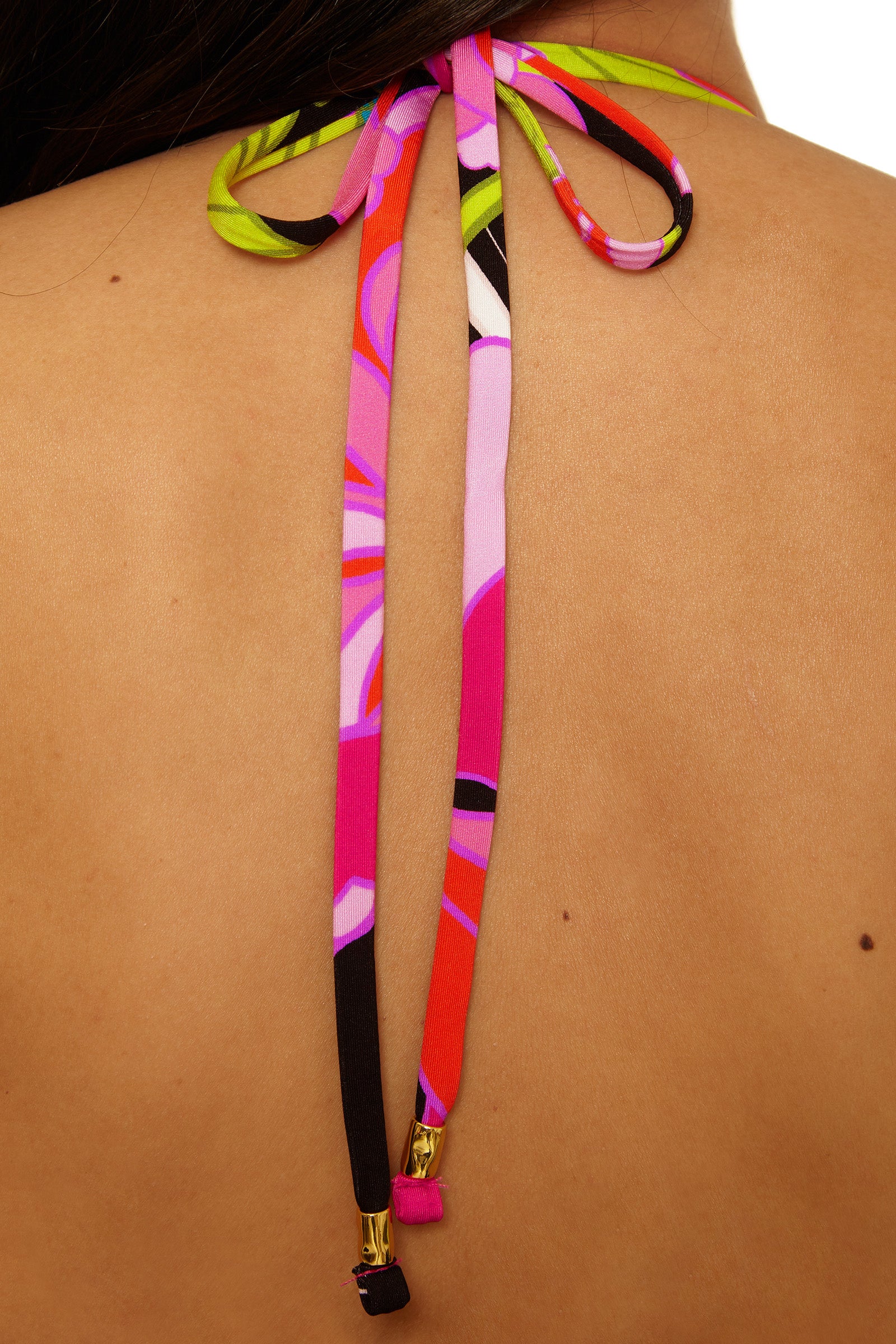 A close-up of a persons back shows the Twist Bandeau Swim Top - Bougainvillea, featuring colorful fabric strings tied in a bow at the neckline. The multicolored strings display pink, green, black, and white patterns with gold tips.