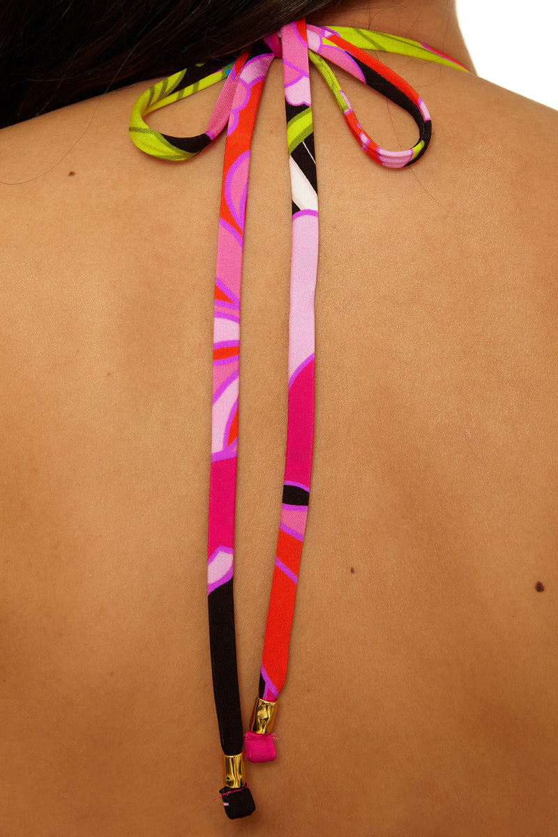 A close-up of a persons back shows the Twist Bandeau Swim Top - Bougainvillea, featuring colorful fabric strings tied in a bow at the neckline. The multicolored strings display pink, green, black, and white patterns with gold tips.