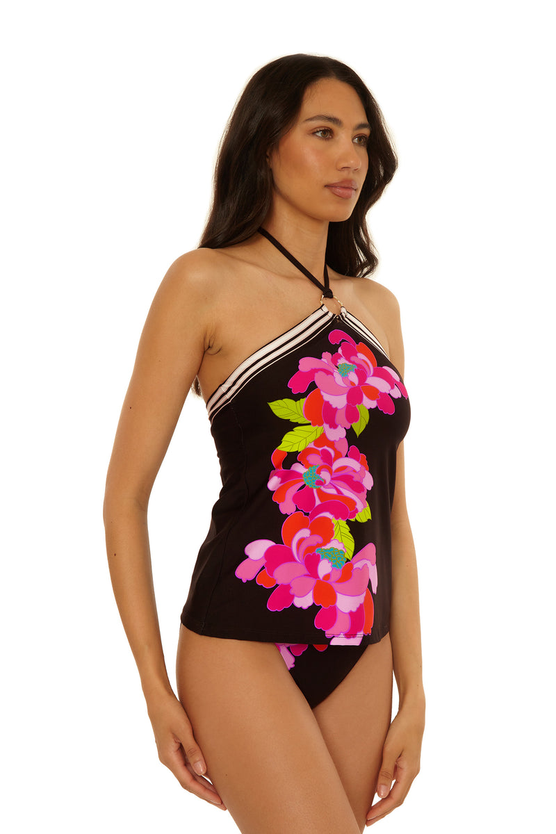 A woman wears the Reversible Tankini Swim Top - Bougainvillea, featuring a black halter-neck style with white trim and a vibrant pink floral pattern, standing against a plain white background.