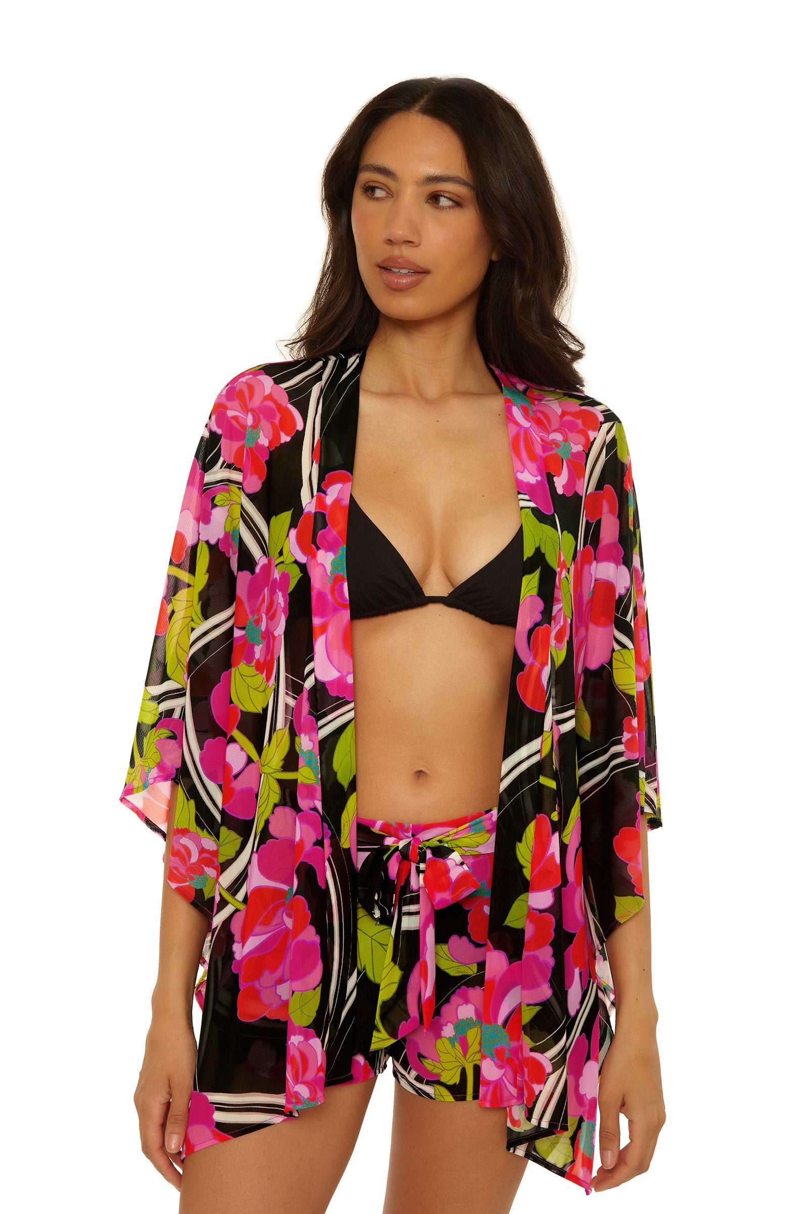 A woman wears the Wrap Cover-Up Short - Bougainvillea set: a black bikini top with a colorful floral kimono and matching shorts in pink, green, and black, posed against a white background.