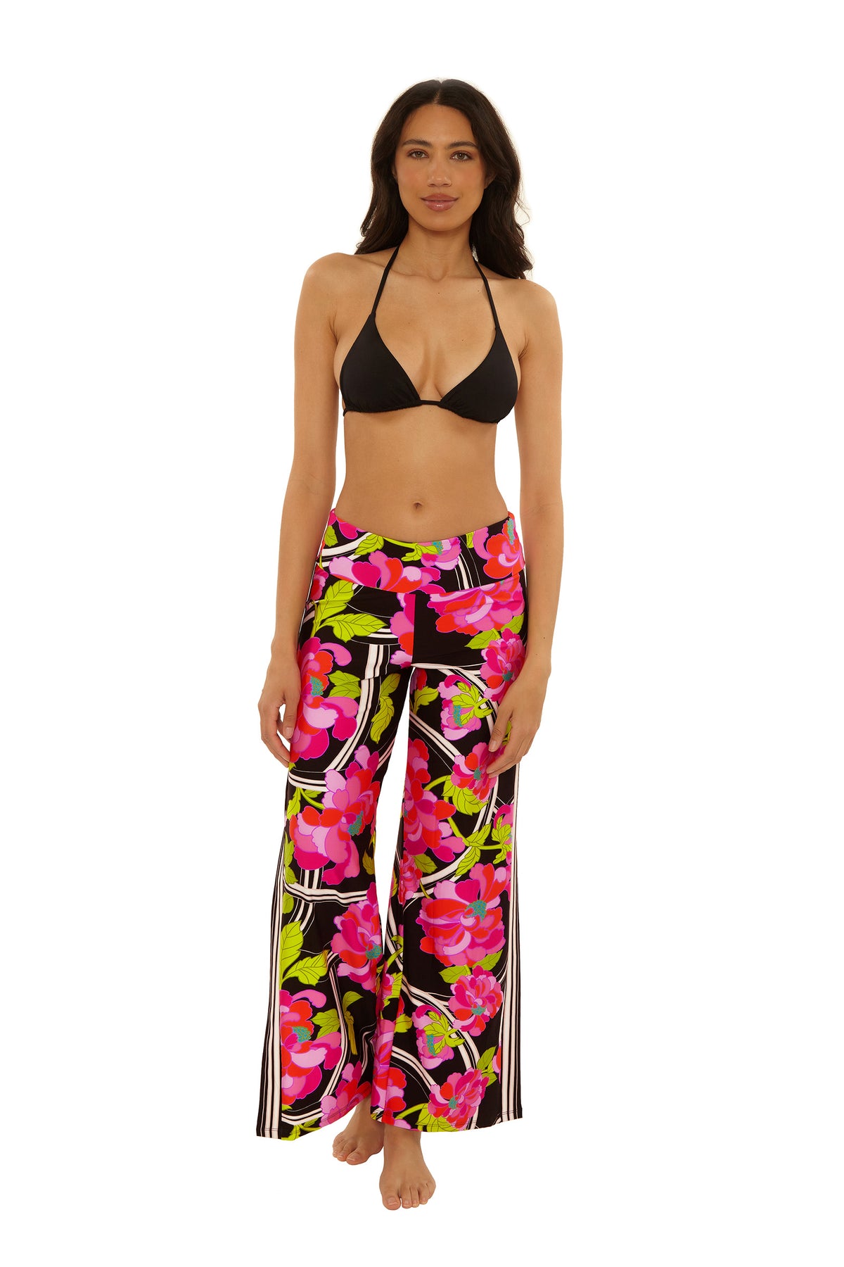 A woman stands barefoot in the Swim Cover-Up Pant - Bougainvillea, featuring a vibrant pink, green, and white floral print on a black background. She pairs it with a black bikini top and poses against a white backdrop.