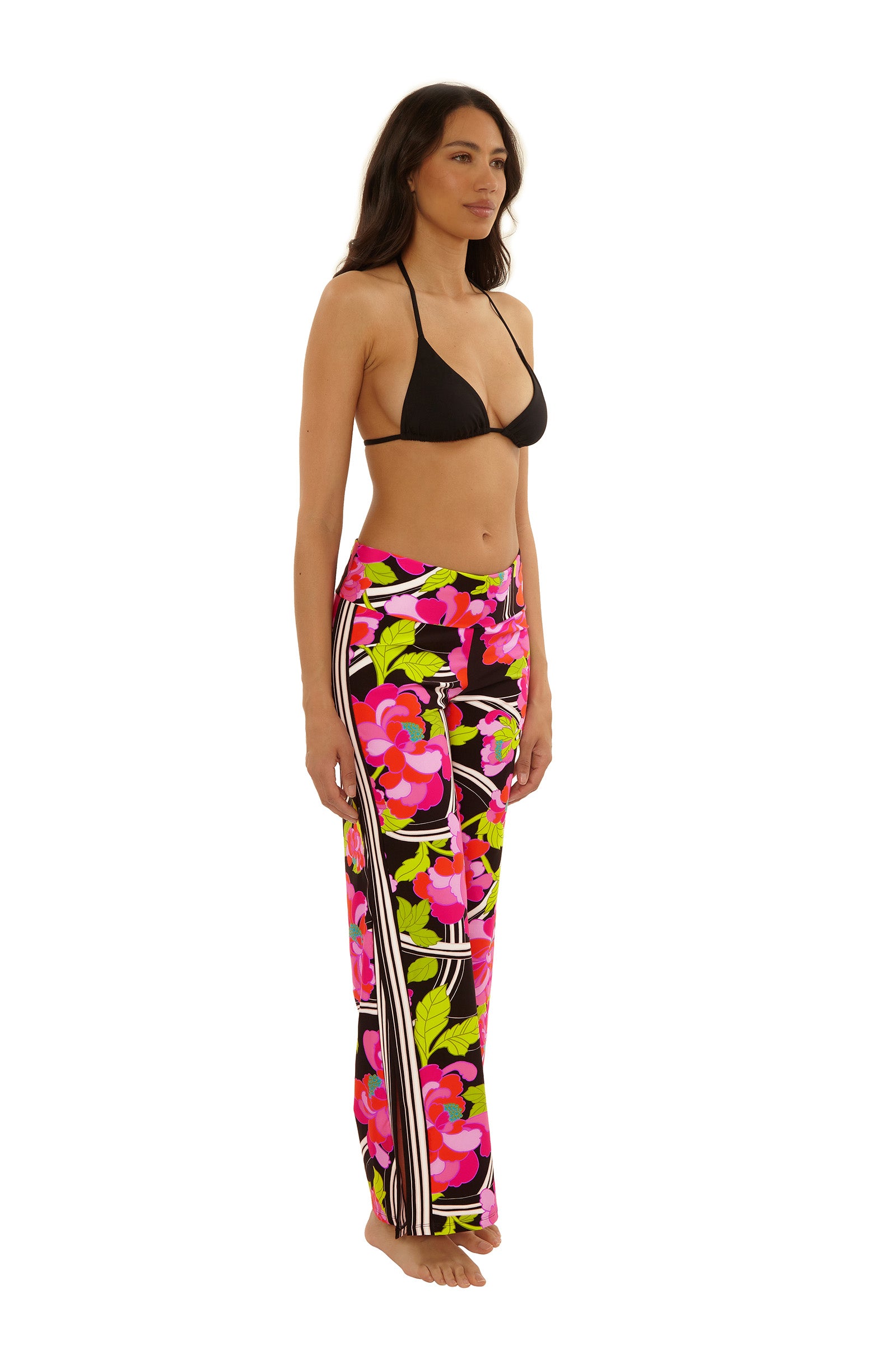 A woman stands barefoot against a plain white background, wearing a black bikini top and the Swim Cover-Up Pant - Bougainvillea, featuring a colorful, high-waisted floral print in pink, red, green, and white.