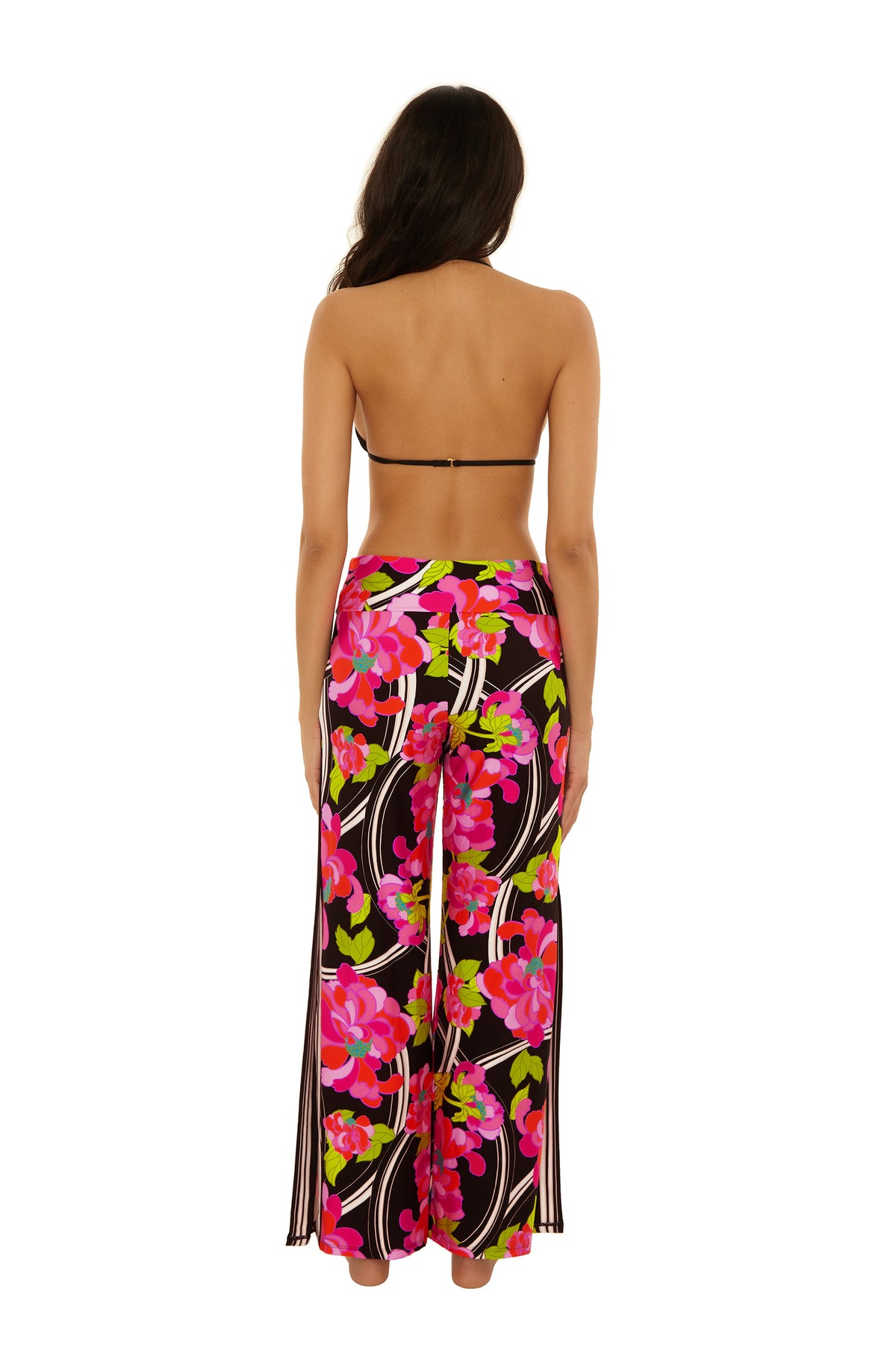 A woman stands barefoot with her back to the camera, wearing a black bikini top and the Swim Cover-Up Pant - Bougainvillea, featuring a vibrant pink, red, and green floral pattern on black.