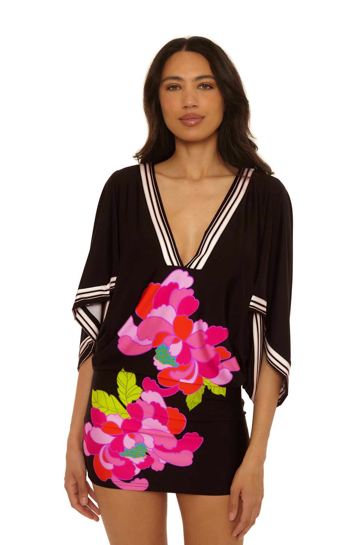 A woman models the Casablanca Tunic Cover-Up in Bougainvillea Swim Knit, featuring wide sleeves, white striped trim, and bold pink and red floral prints with green leaves, against a white background.