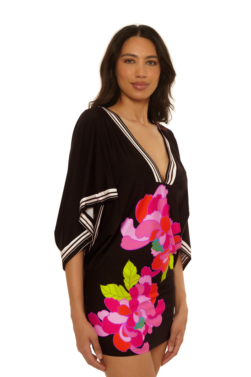 A woman wears the Casablanca Tunic Cover-Up in Bougainvillea, featuring wide sleeves and a vibrant pink, red, and yellow floral print, standing against a plain white background.