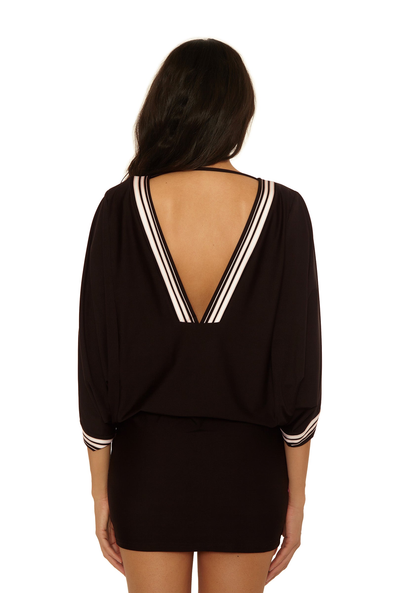 A woman faces away, wearing the Casablanca Tunic Cover-Up in Bougainvillea—a black dress featuring a deep V-shaped back and white striped trim on the neckline and sleeves. Her long dark hair is down against a white background.