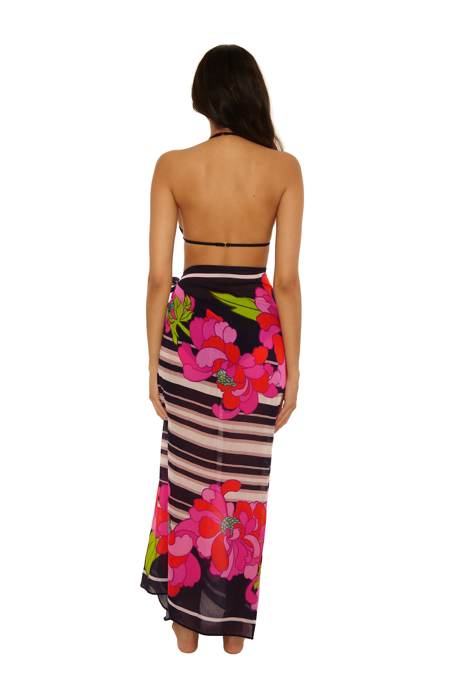 A woman stands with her back to the camera, wearing a halter-top black bikini and the Pareo Swim Cover-Up - Bougainvillea, a long, sheer sarong with bold pink floral and horizontal stripe patterns against a plain white background.