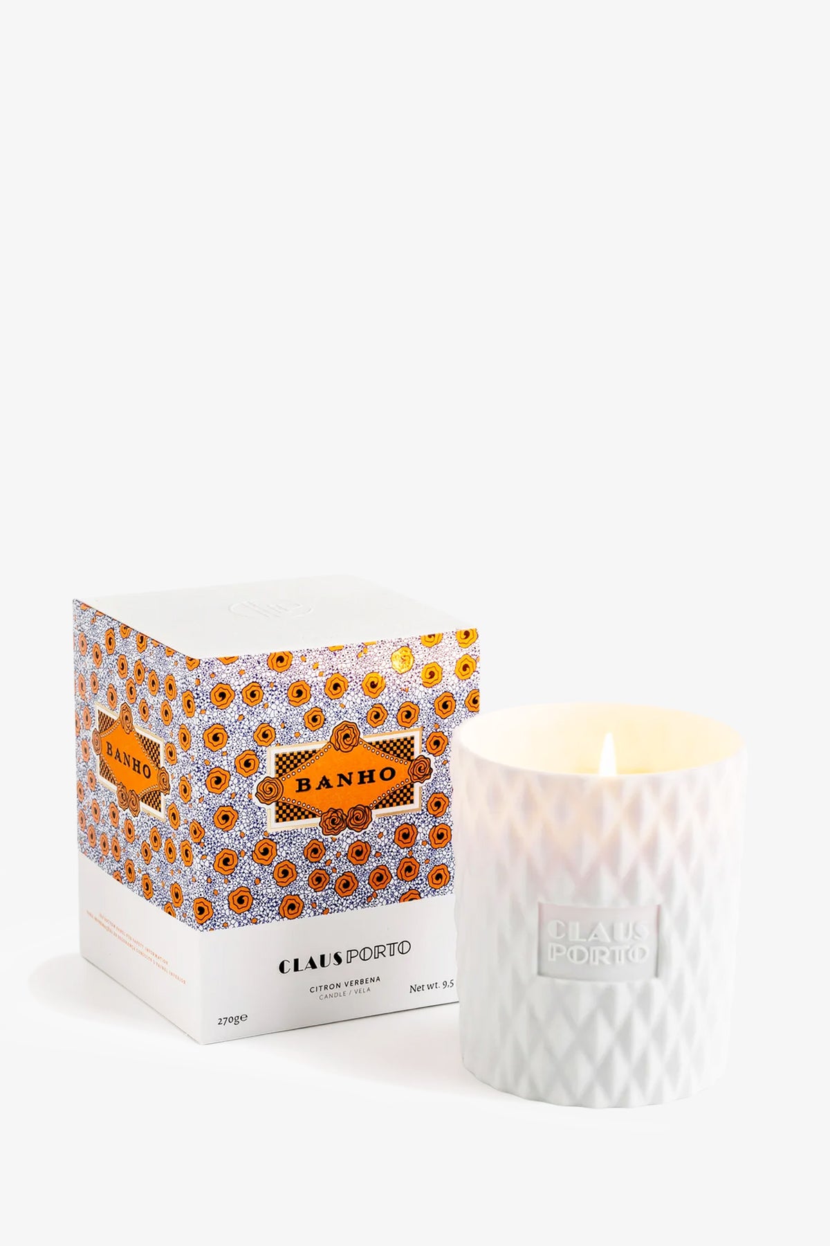 A Claus Porto Banho Candle featuring a white wax in a textured glass holder is placed next to its orange-patterned box against a plain white background.