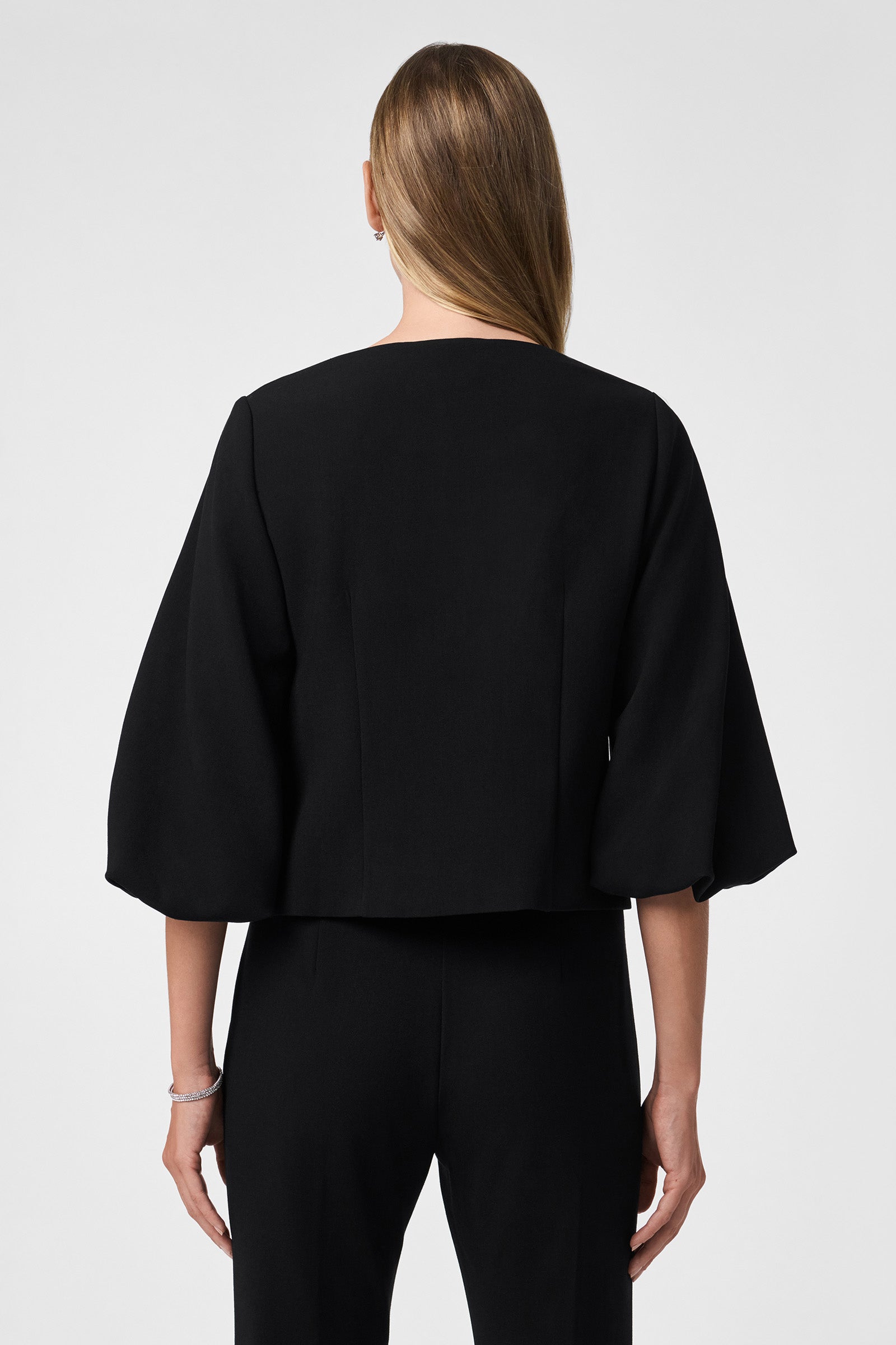 A woman with straight, light brown hair models the Basal Cropped Jacket—loose-fitting with wide three-quarter sleeves—in black, paired with matching pants, facing away from the camera against a plain light background.