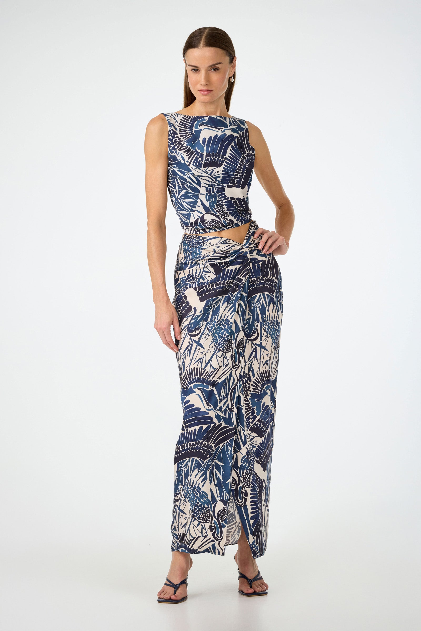 A woman stands confidently in the Hakuto Tie-Back Top - Tsuru, a sleeveless blue and white tropical print top with side cutouts, paired with black sandals, against a plain white background.