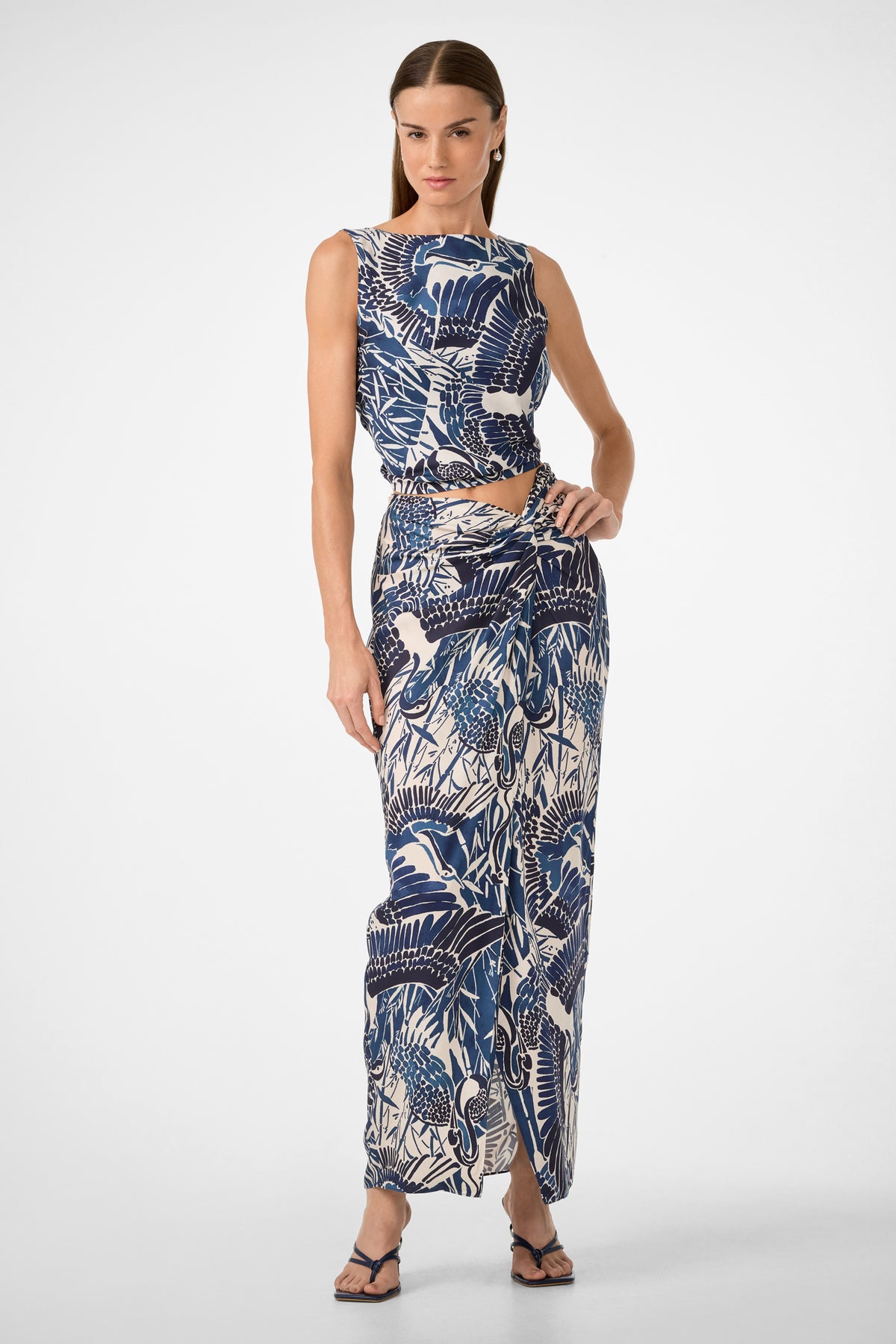 A woman models the Alodia Tie-Front Maxi Skirt - Tsuru, a blue and white printed, high-waisted, ankle-length skirt with a side slit. She pairs it with a matching sleeveless top and black sandals against a plain white background.
