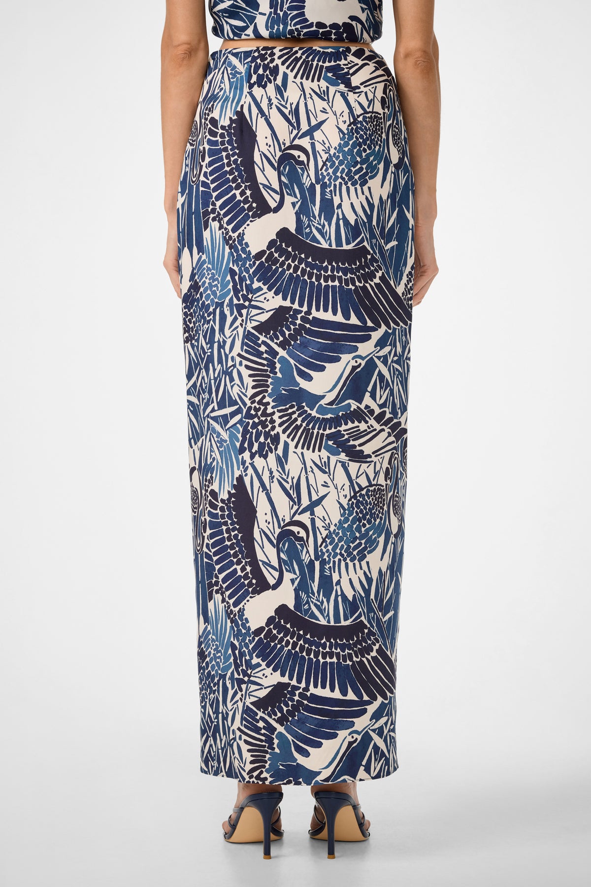 A woman is seen from the back wearing the Alodia Tie-Front Maxi Skirt - Tsuru, featuring a blue and white bird and foliage print, paired with dark heeled sandals against a plain white background.
