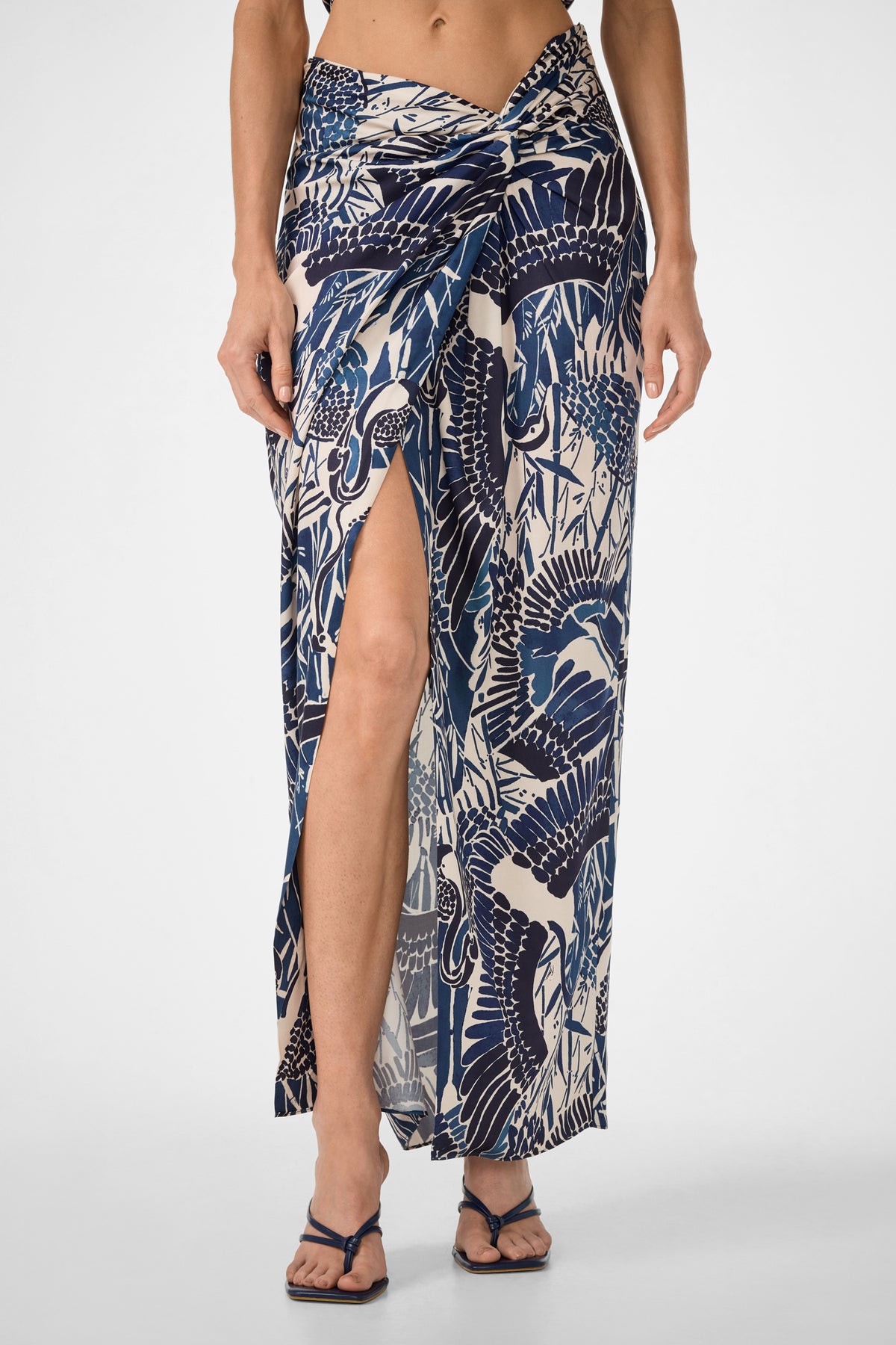 A woman wears the Alodia Tie-Front Maxi Skirt in Tsuru, featuring a long patterned wrap design with a front slit that reveals one leg, paired with dark flip-flops. The photo is cropped from mid-torso to feet.