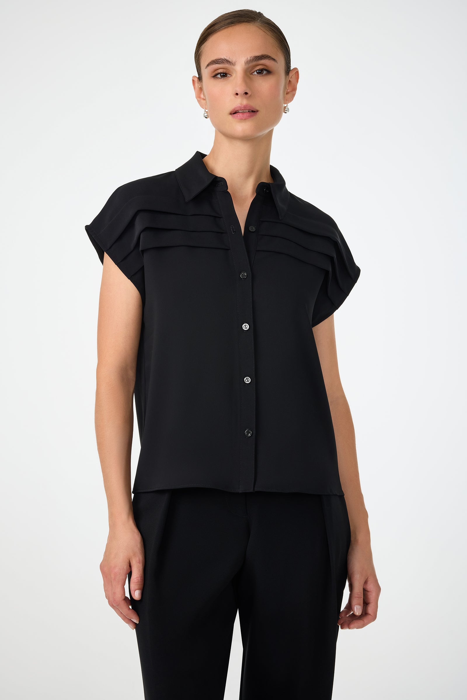 A woman wears the Cheri Pleated Button-Front Top in black, paired with black trousers, standing against a plain light background.