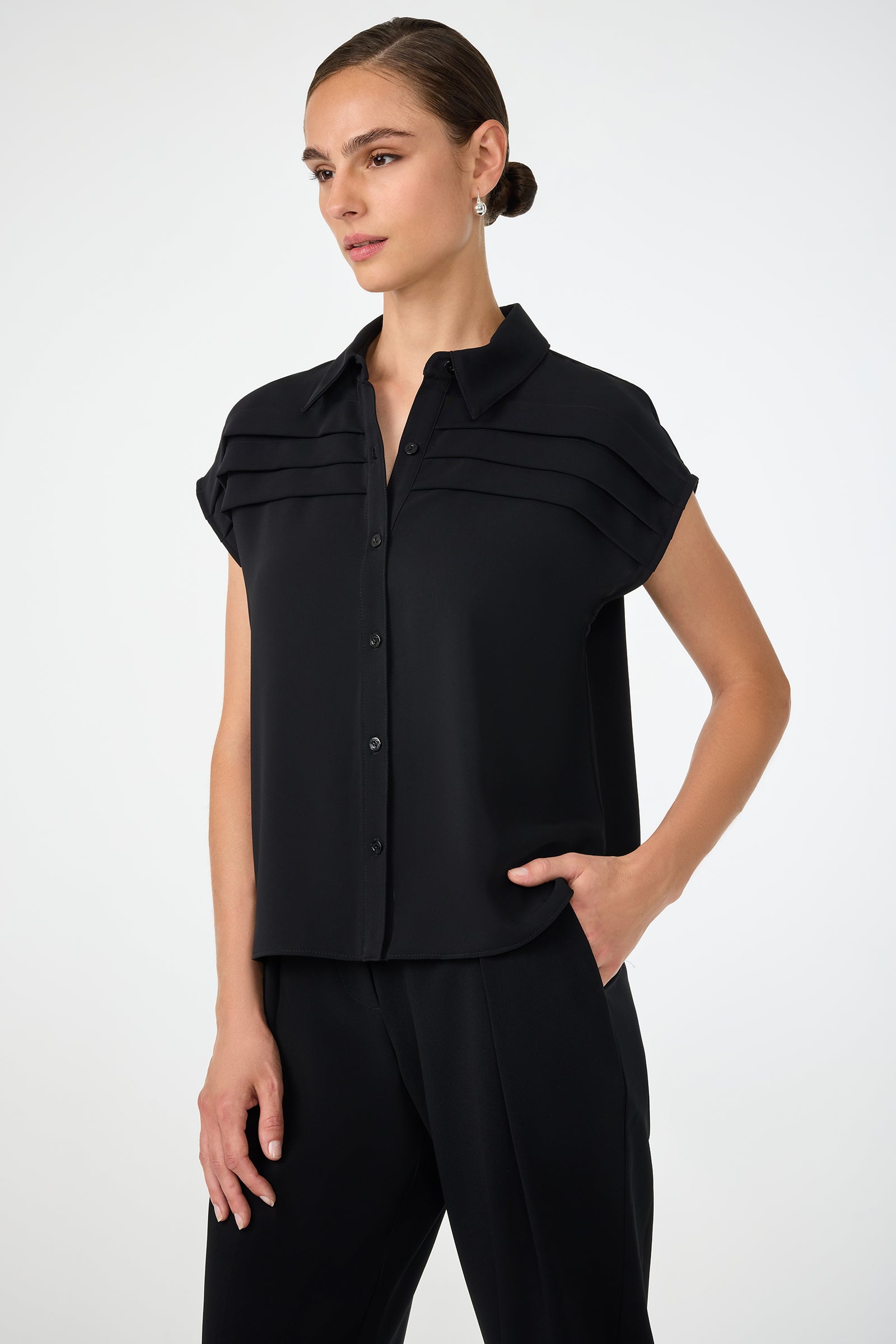 A woman wearing the Cheri Pleated Button-Front Top, a short-sleeve black blouse with horizontal chest pleats, and matching black pants stands with one hand in her pocket against a plain white background.