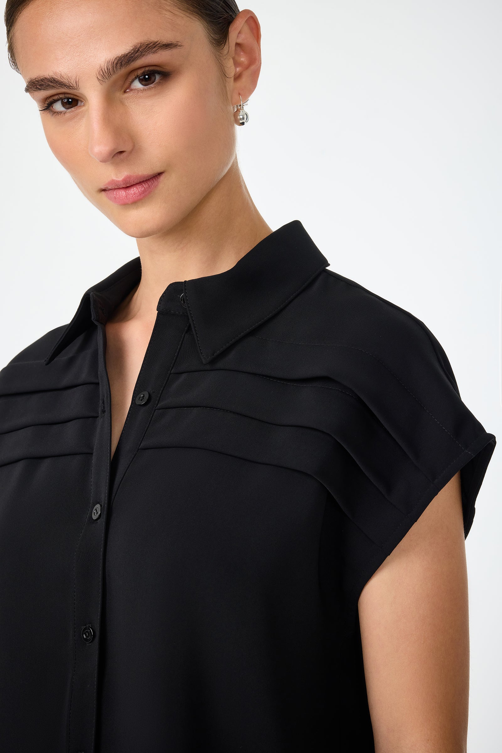 A woman with brown hair pulled back wears the Cheri Pleated Button-Front Top, a black short-sleeved blouse with layered shoulder detailing, looking confidently at the camera against a plain background.