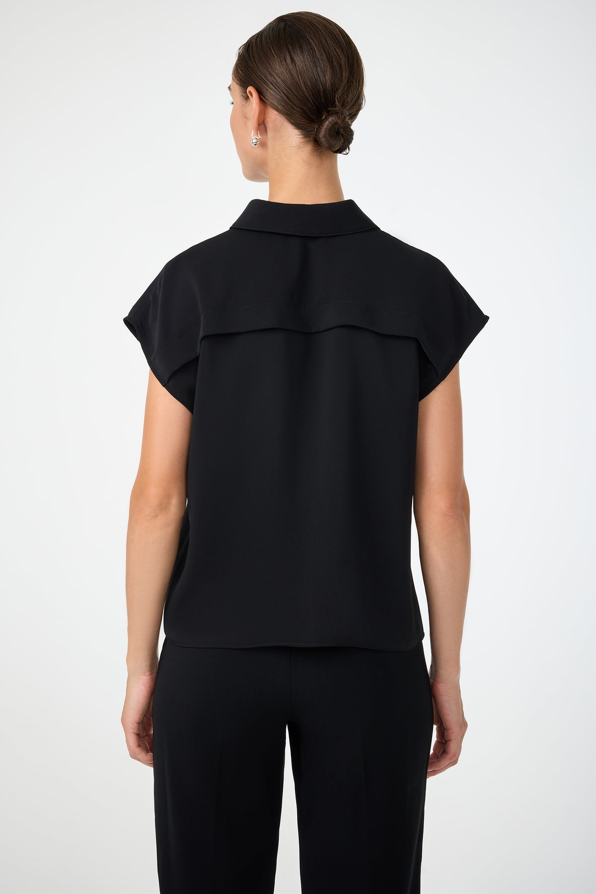 A woman stands facing away, wearing the Cheri Pleated Button-Front Top in black with matching pants. Her hair is styled in a neat bun, and she poses against a plain, light-colored background.