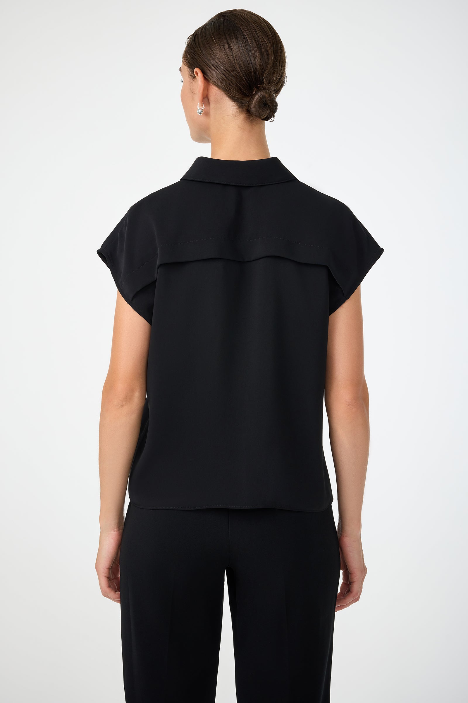 A woman stands facing away, wearing the Cheri Pleated Button-Front Top in black with matching pants. Her hair is styled in a neat bun, and she poses against a plain, light-colored background.