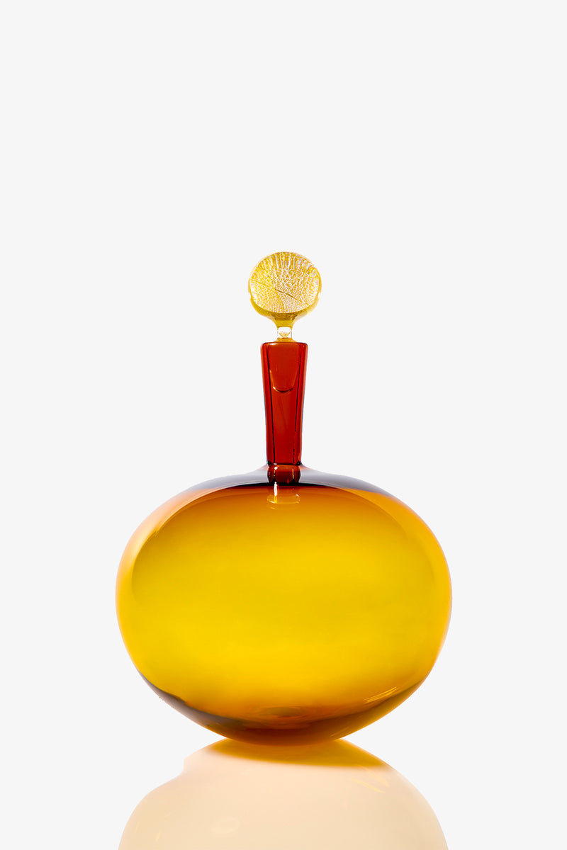 The Carmella Barware Low Round is a round, amber glass bottle with a smooth surface, a short red neck, and a decorative yellow seashell-shaped stopper, shown on a plain white background.