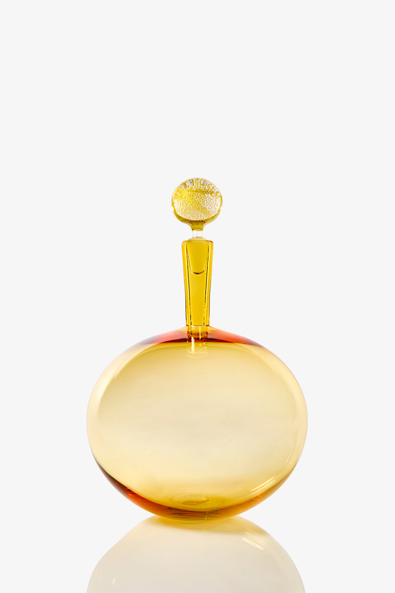 The Carmella Barware Low Round is a round, golden-yellow glass bottle with a flat base and a tall, narrow stopper topped with a textured circular design, displayed against a plain white background.