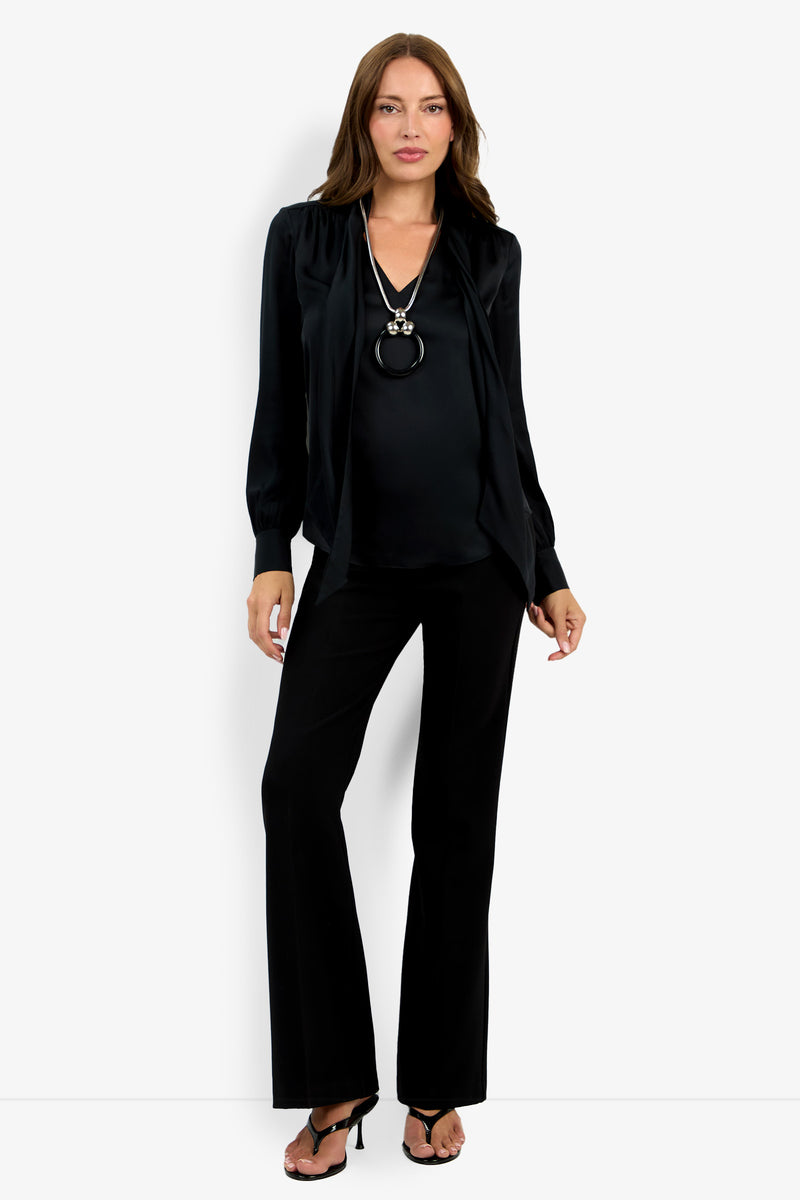 A woman with straight brown hair and a neutral expression stands against a light background wearing the Deming Top, black wide-leg pants, heeled sandals, and a long silver necklace.