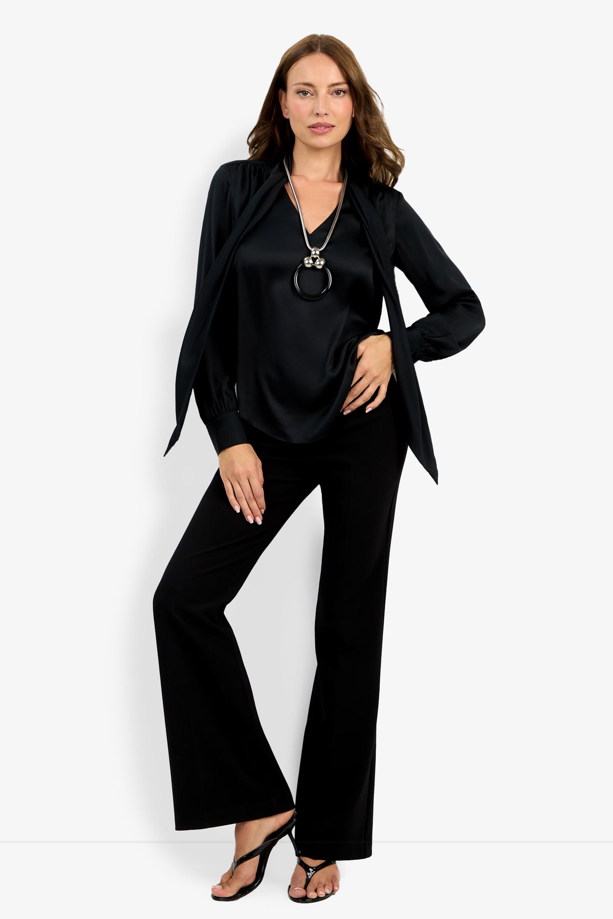A woman poses against a white background wearing the Deming Top, black flared pants, black heeled sandals, and a long pendant necklace, with one hand on her hip and the other resting on her thigh.
