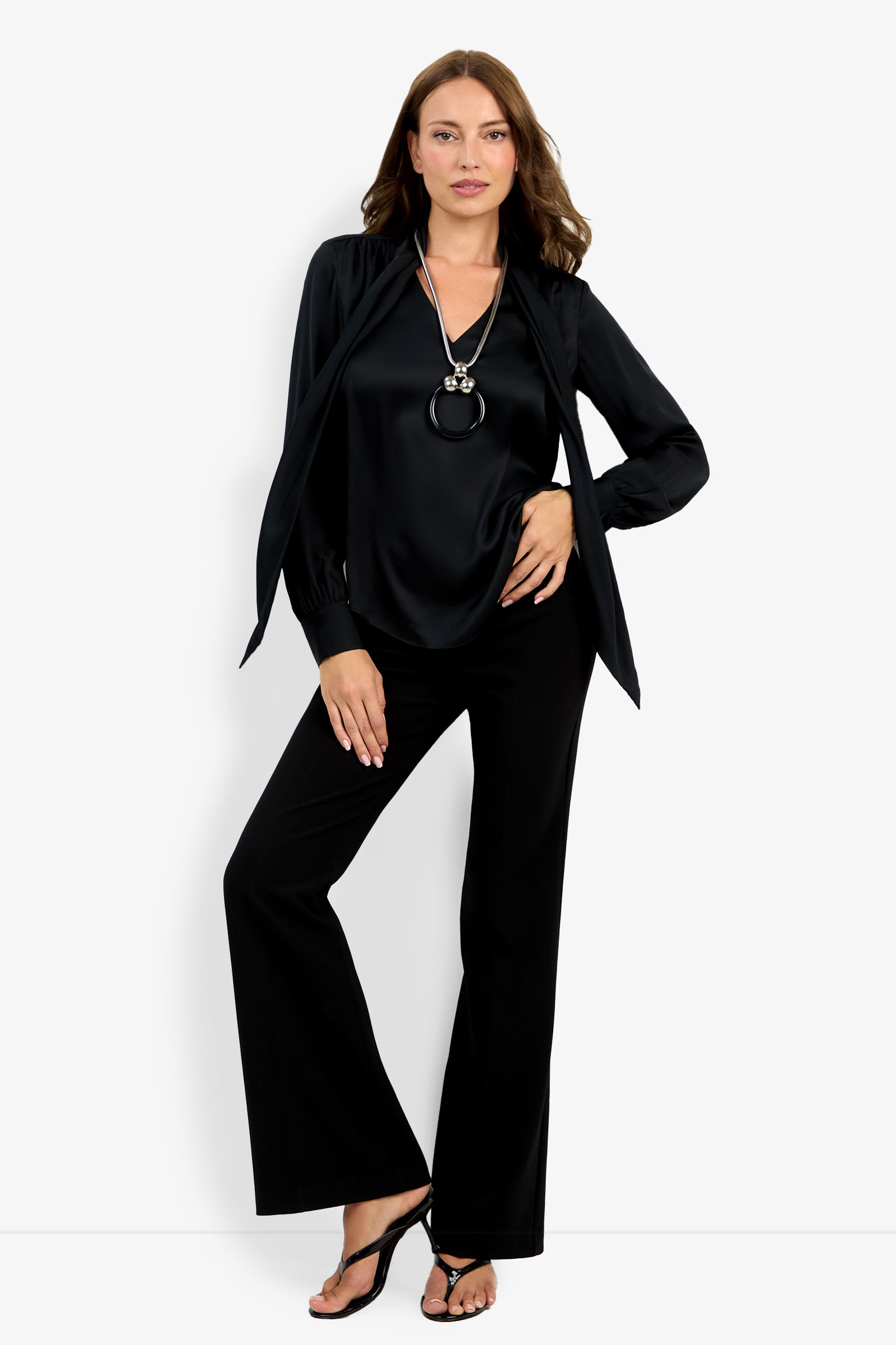 A woman poses against a white background wearing the Deming Top, black flared pants, black heeled sandals, and a long pendant necklace, with one hand on her hip and the other resting on her thigh.