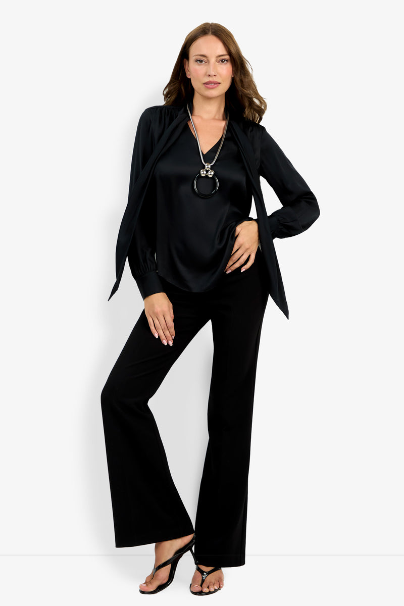 A woman poses against a white background wearing the Deming Top, black flared pants, black heeled sandals, and a long pendant necklace, with one hand on her hip and the other resting on her thigh.