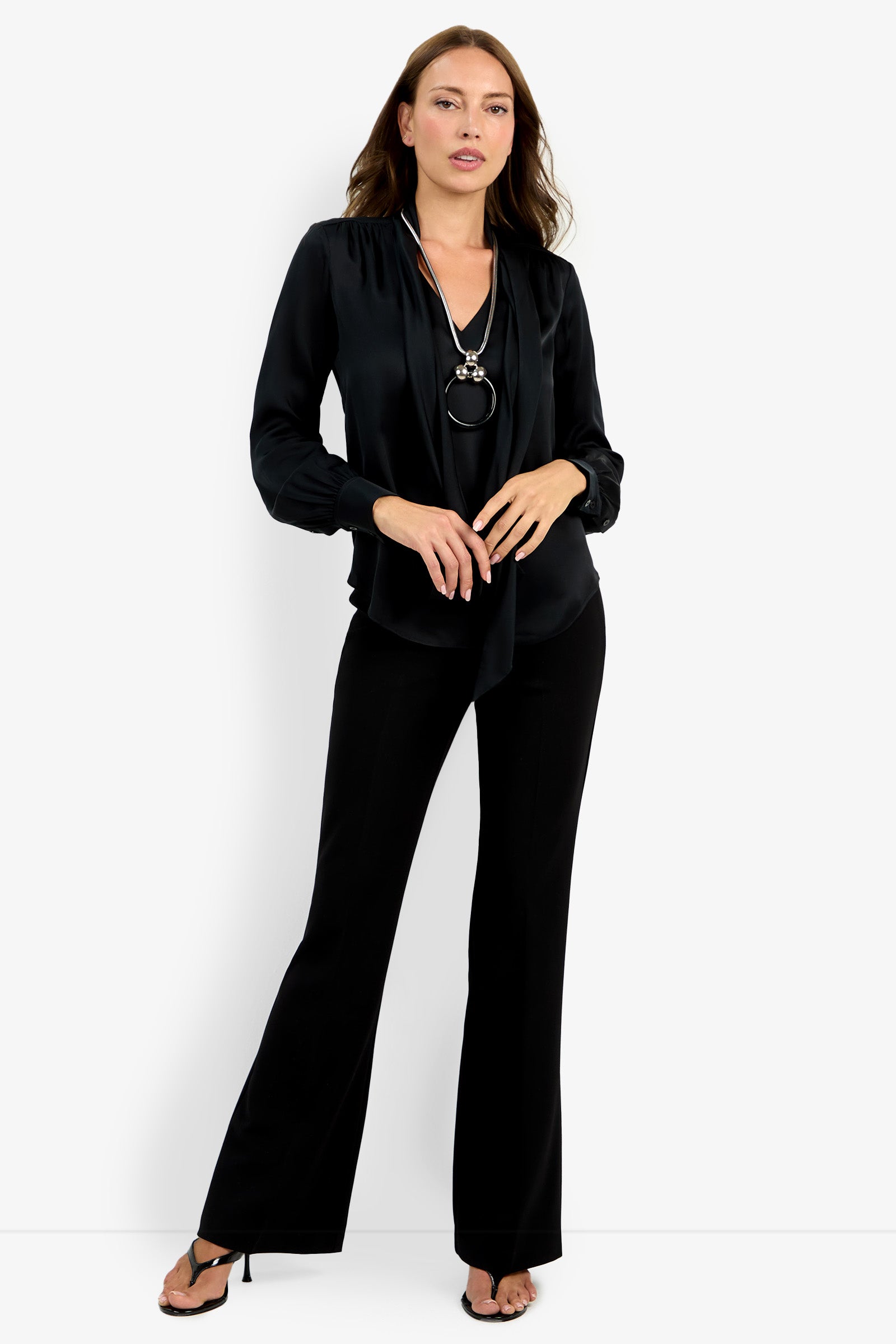 A woman stands against a white background wearing the Deming Top, a black long-sleeve blouse with a loose tie detail, paired with black flared pants, black high-heeled sandals, and a statement necklace.