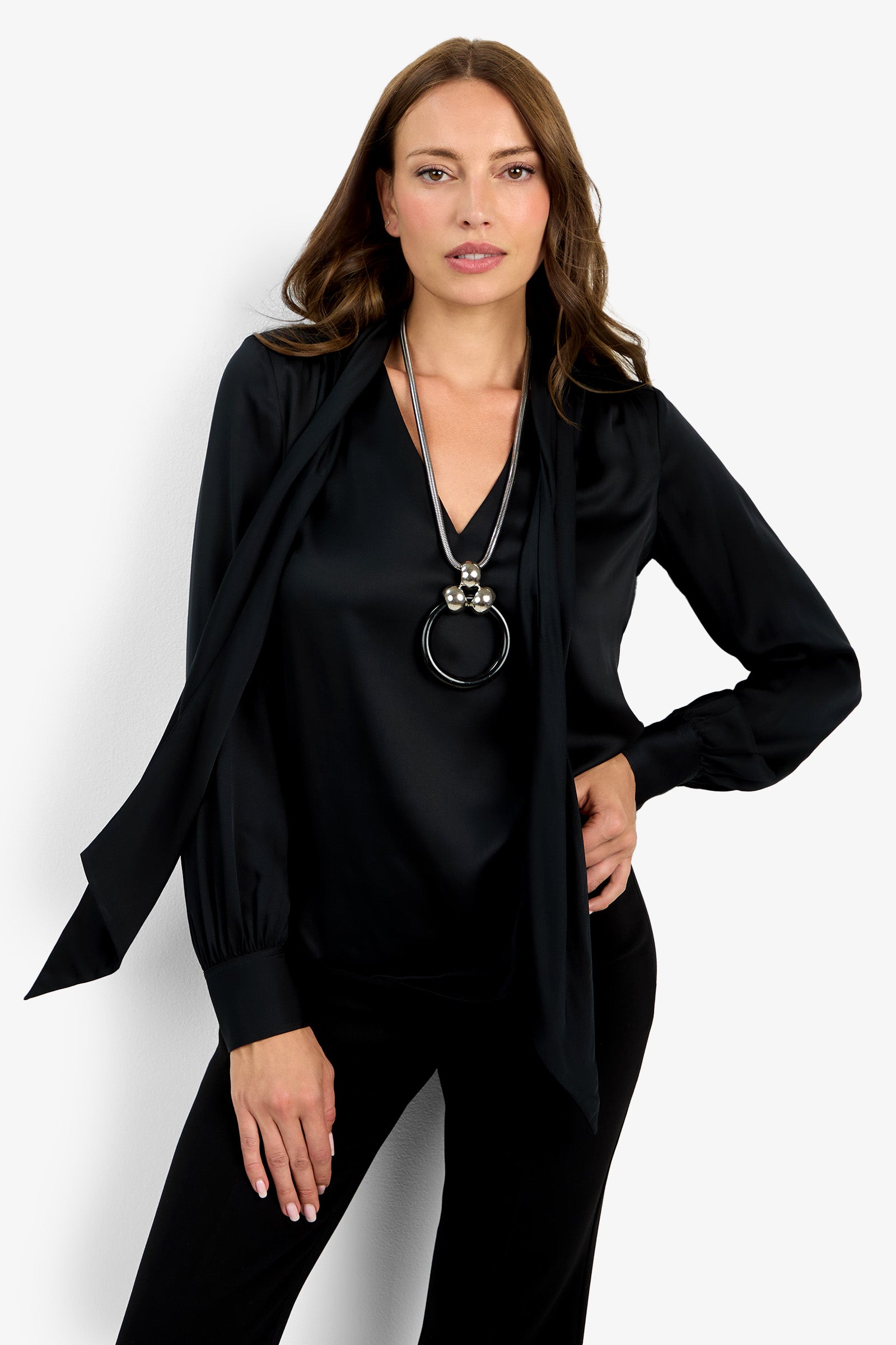 A woman stands confidently with one hand on her hip against a white background, wearing the Deming Top—a black long-sleeve blouse with a necktie detail—paired with a large pendant necklace.