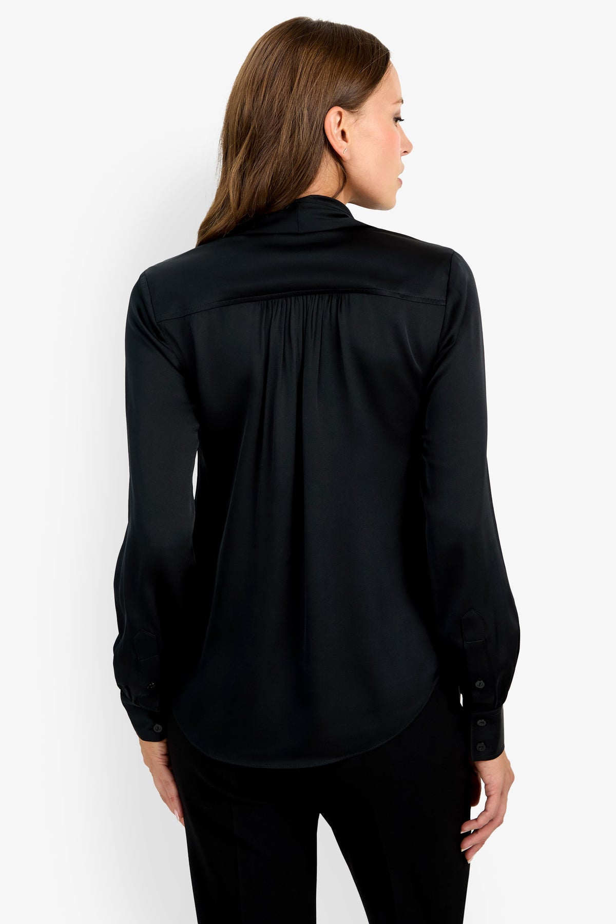 A woman with straight brown hair is wearing the Deming Top, a long-sleeve black blouse, and black pants. She stands with her back to the camera, looking over her shoulder to the right against a plain white background.