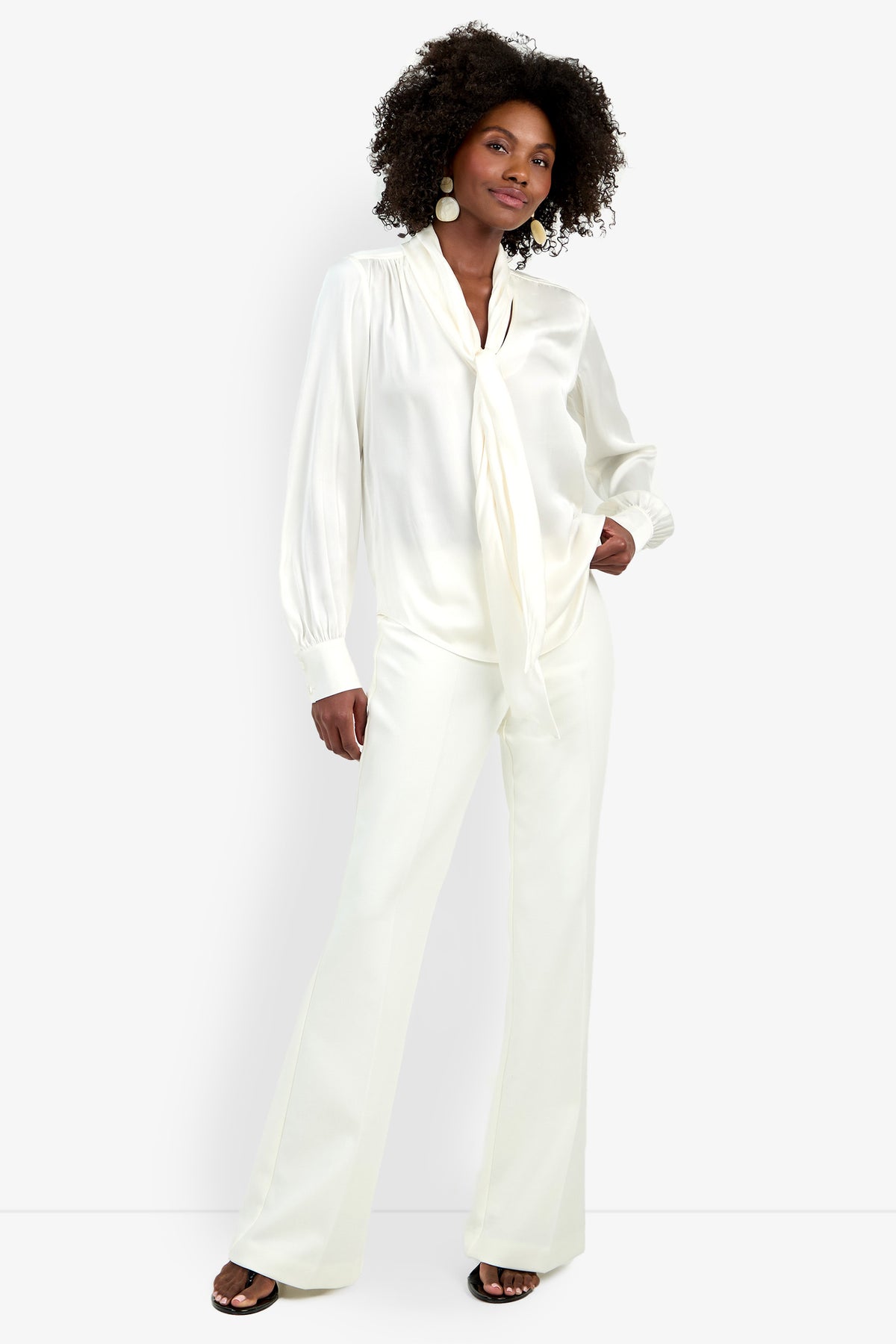 A woman stands confidently in the Deming Top, a silky white blouse with a tie-neck detail, paired with matching wide-leg trousers. She has natural curly hair, large round earrings, and black open-toe shoes against a plain background.