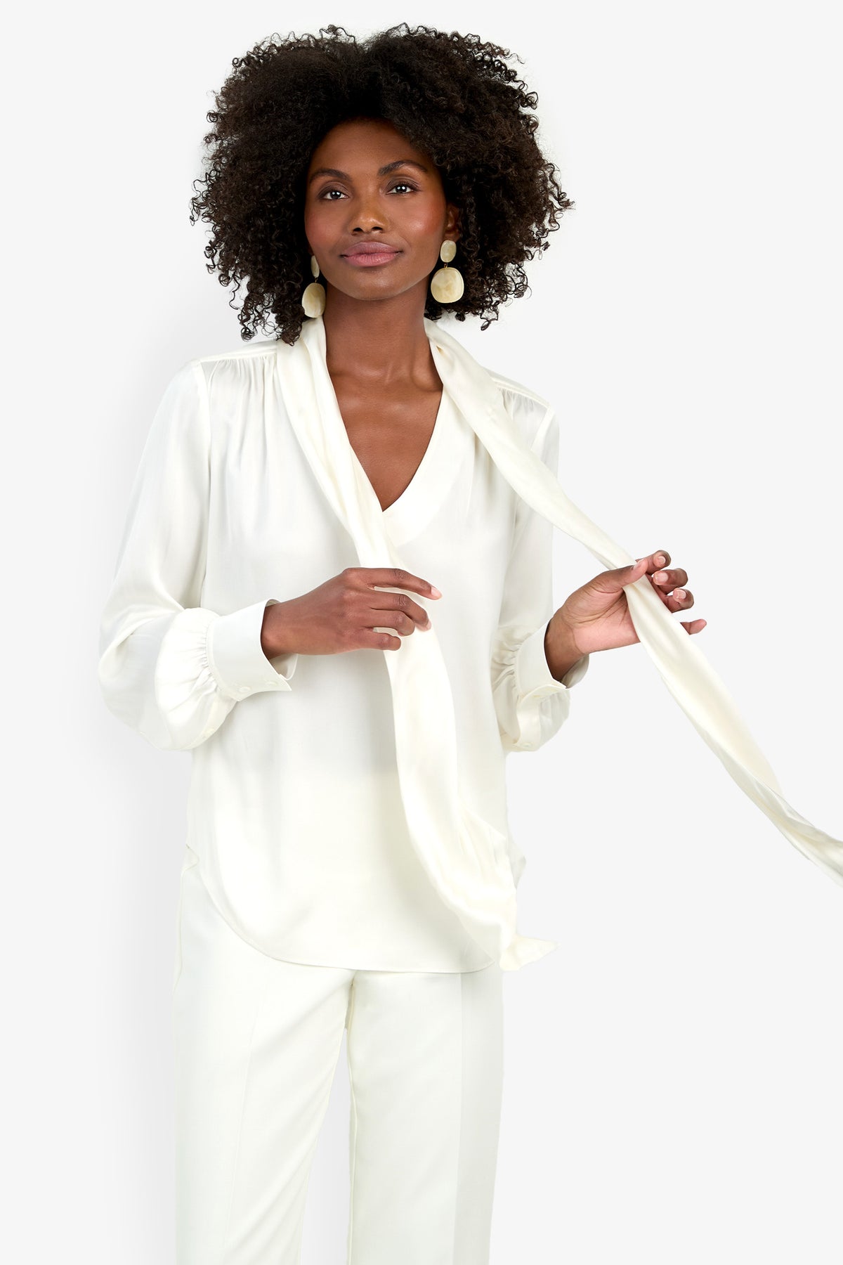 A woman with curly hair wears the Deming Top, a white blouse with a loose tie detail, and white pants. She stands against a plain white background, holding one end of the tie and smiling gently.