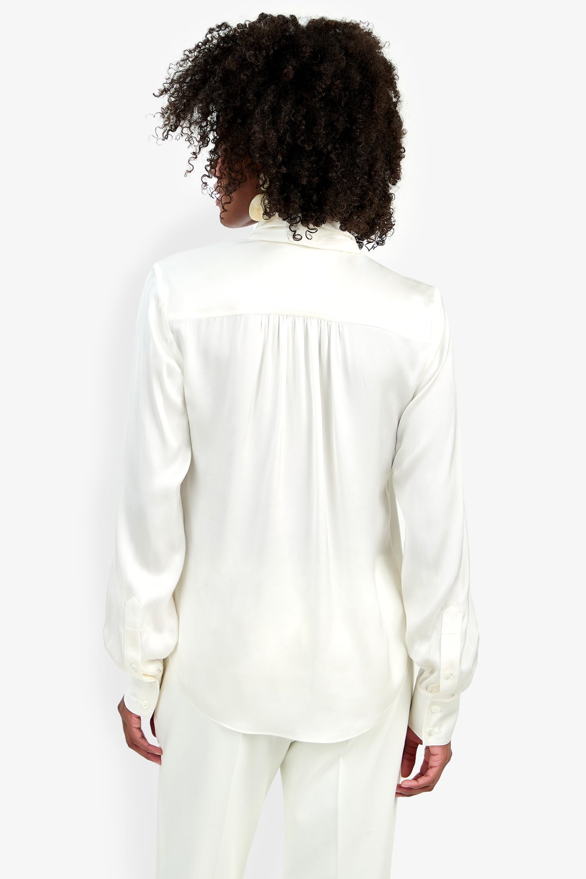 A person with curly hair is seen from behind, wearing the Deming Top—a long-sleeve white blouse—paired with matching white pants, standing against a plain white background.