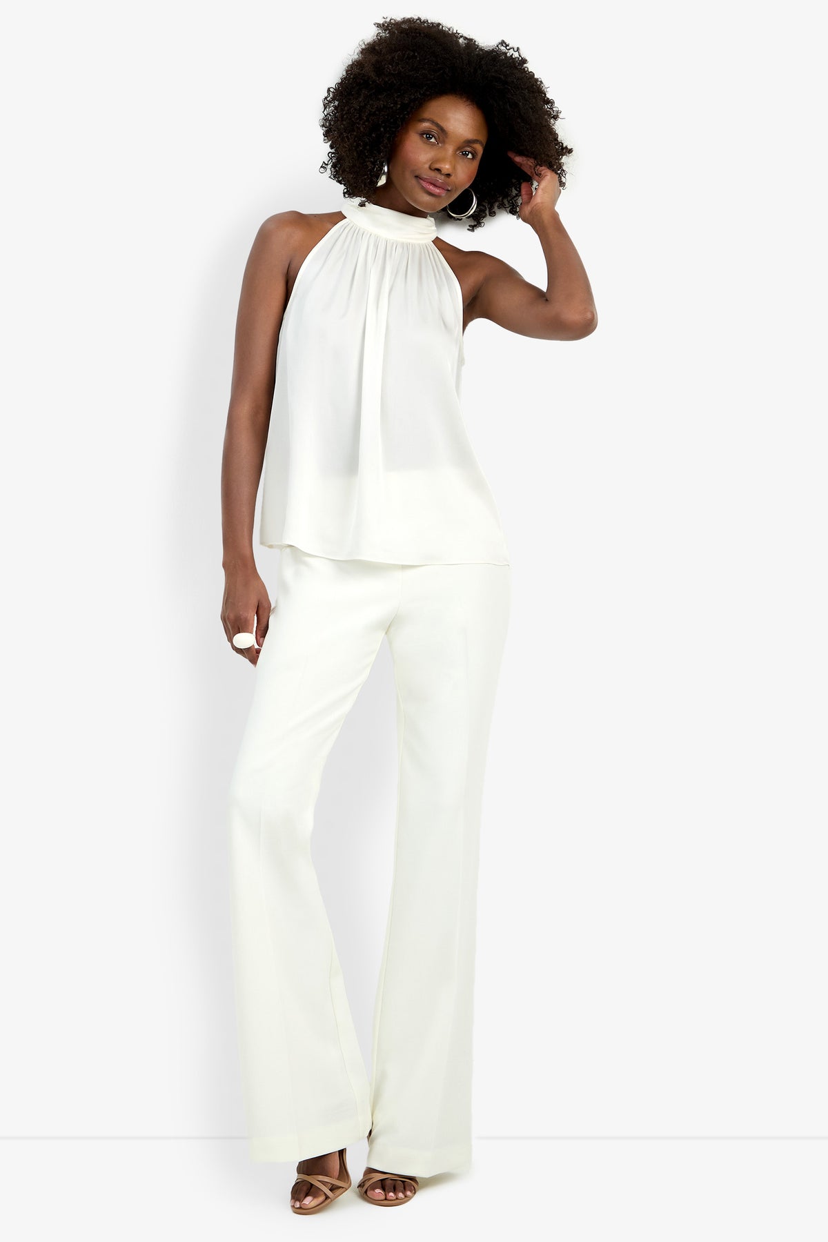 A woman with curly hair confidently wears the Zana Top, a sleeveless halter-neck white top, paired with white wide-leg pants and beige heels against a light background.