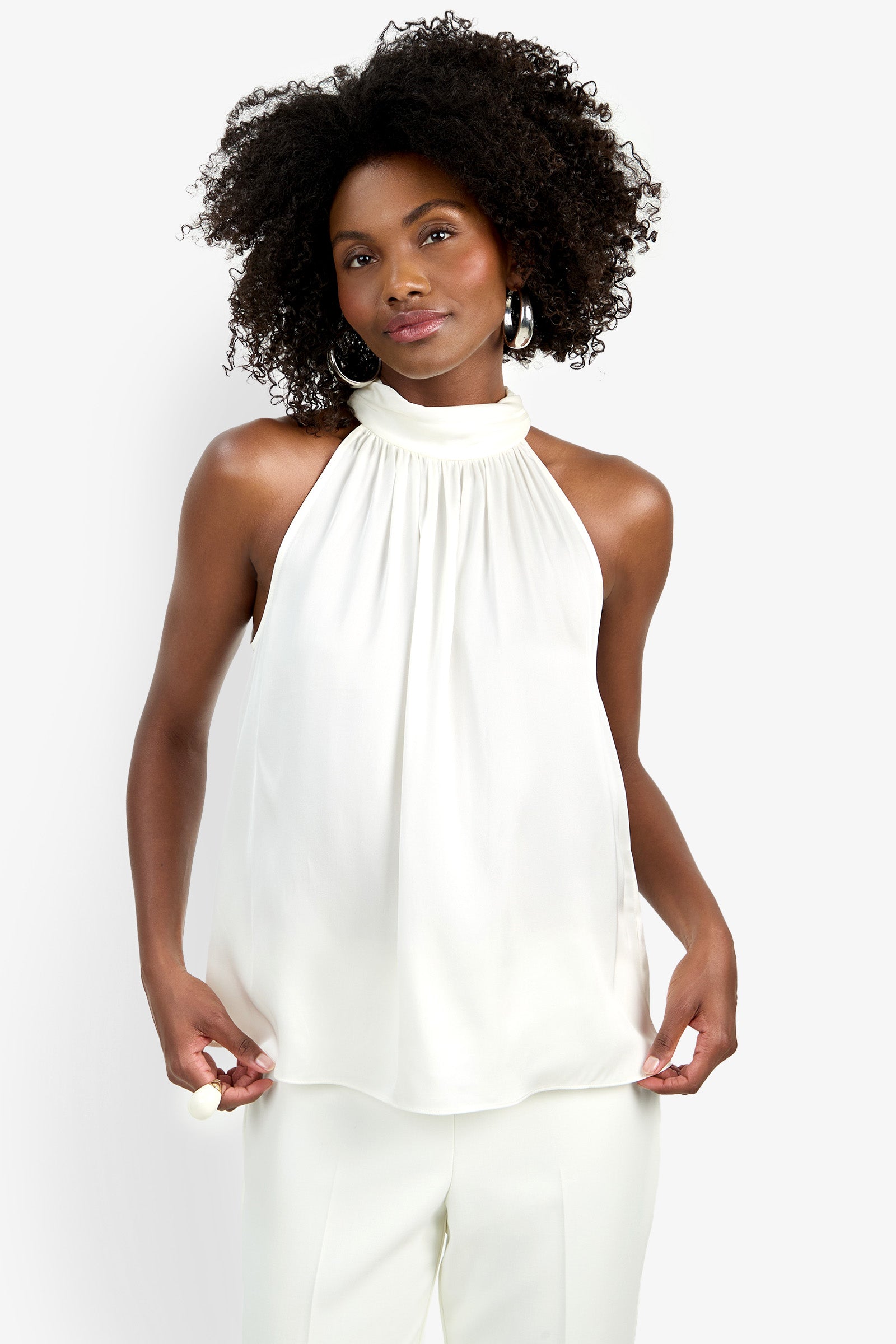 A woman with curly hair wears the sleeveless, high-neck, white Zana Top and white pants. She stands against a plain light background, looking at the camera with a neutral expression and holding the sides of her blouse.