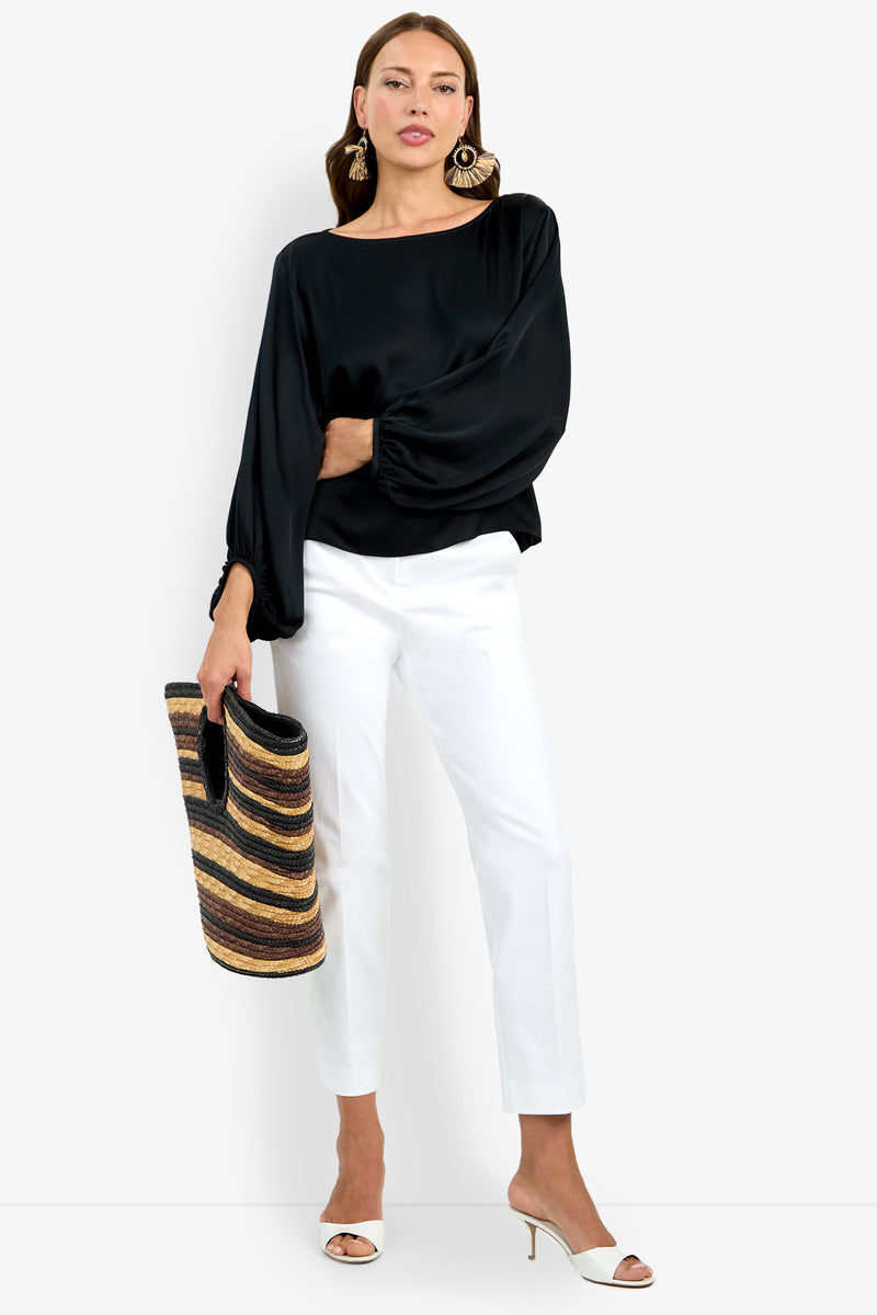 A woman wearing the Ursa Top, black long-sleeve blouse, white pants, and white heeled sandals holds a striped woven tote bag. She accessorizes with large statement earrings and stands against a plain background.