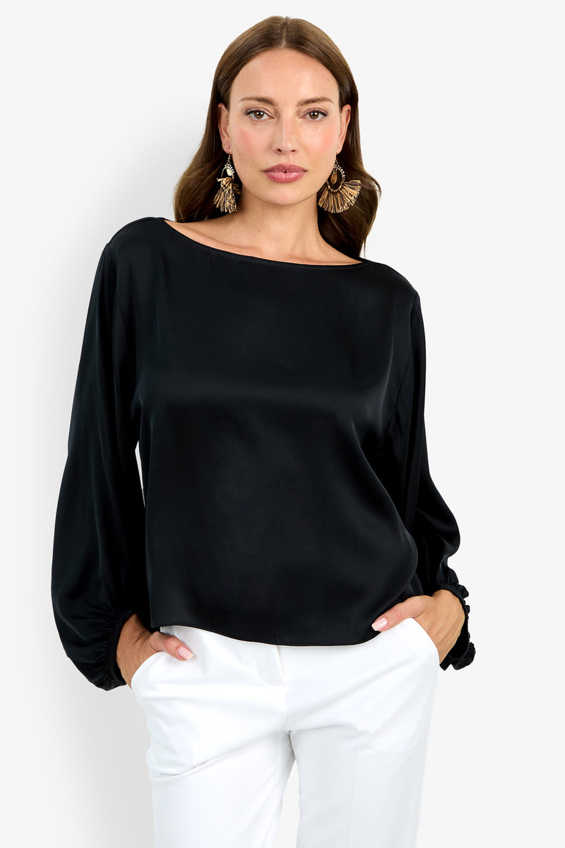 Woman with long brown hair wears statement earrings, white pants, and the Ursa Top—a loose-fitting black satin blouse with long sleeves—standing hands in pockets against a plain white background.