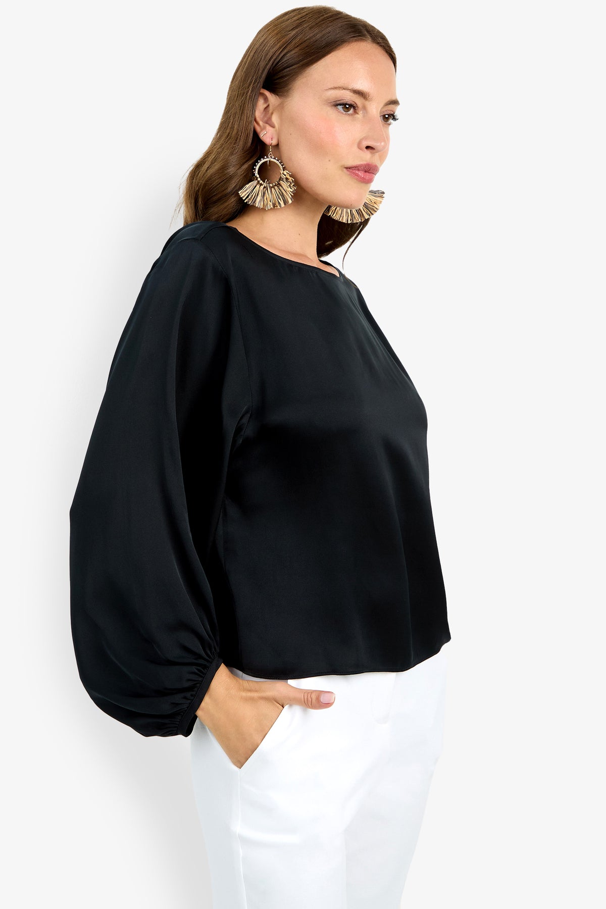 A woman with long brown hair and statement earrings stands sideways with one hand in her pocket, wearing the Ursa Top—a loose black long-sleeve blouse with balloon sleeves—paired with white pants.