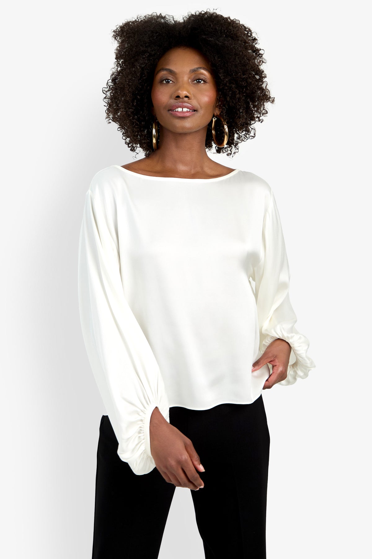 A woman with curly hair wears gold hoop earrings, black pants, and the Ursa Top—a loose-fitting, long-sleeved white blouse with gathered cuffs. She stands against a plain white background, smiling slightly.
