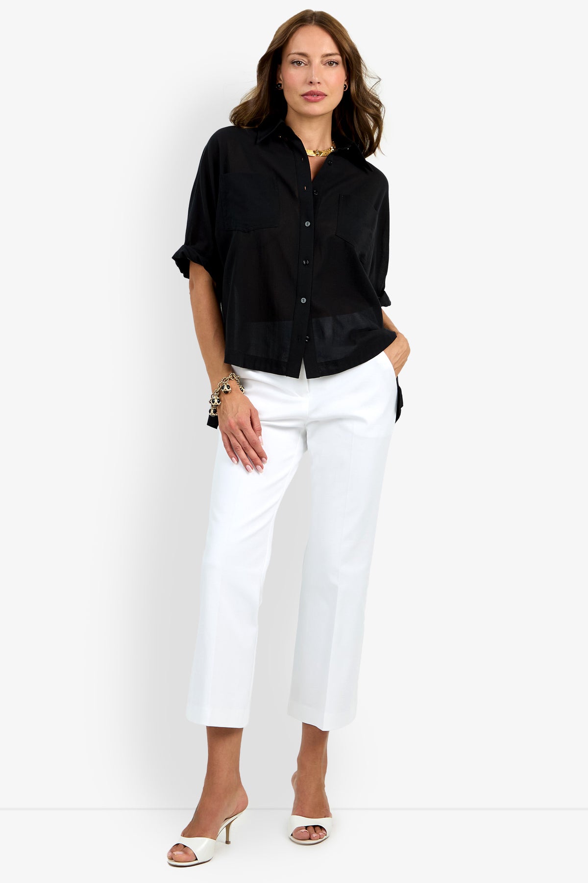 A woman with wavy brown hair wears the Hawaii Top, a loose black button-up shirt, paired with white cropped pants and white heeled sandals. She accessorizes with gold jewelry and has her hands in her pockets.