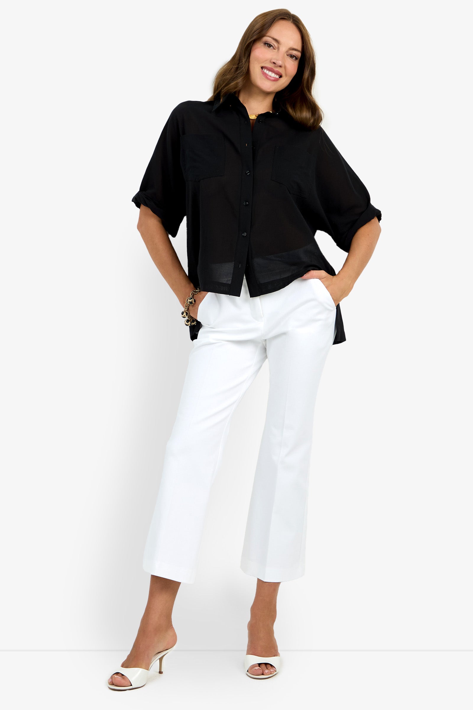 A woman smiles while posing in the Hawaii Top—a loose black button-up shirt with rolled sleeves—paired with white cropped pants and heeled sandals against a plain white background.