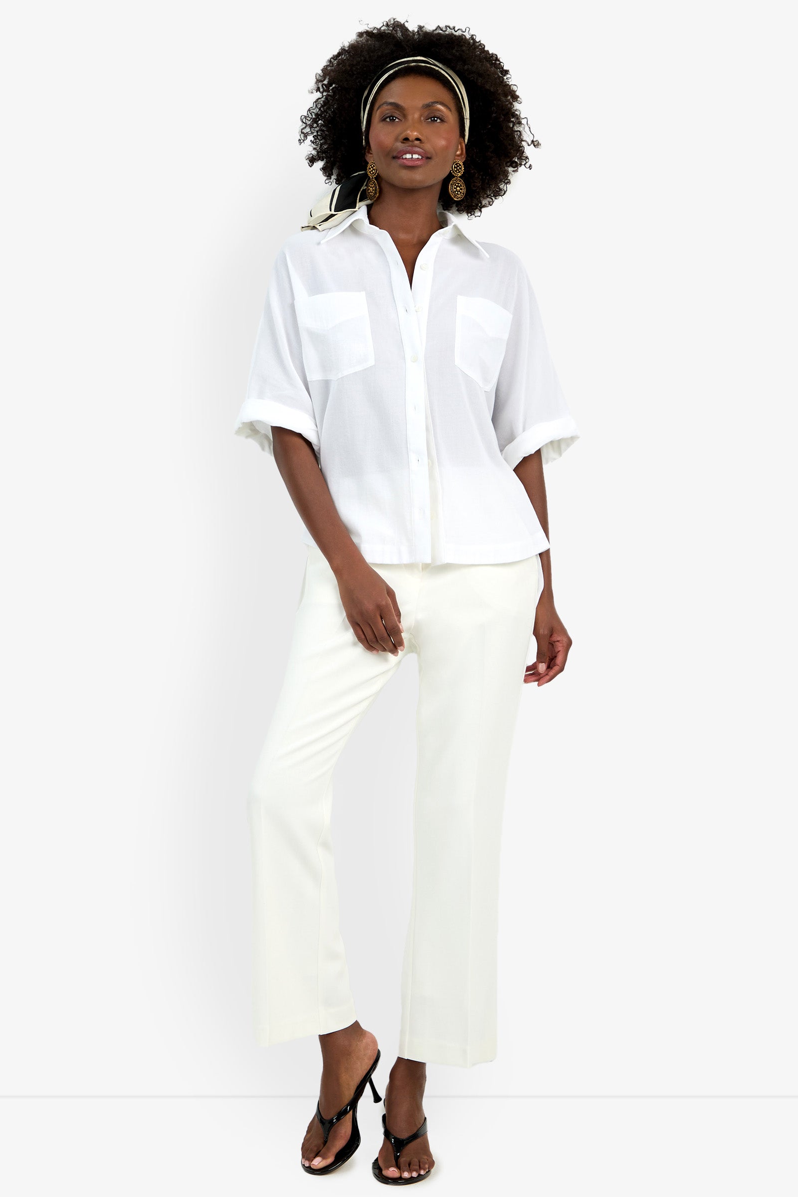 A woman stands confidently against a plain white background wearing the Hawaii Top, white pants, black sandals, gold earrings, and a patterned headband.